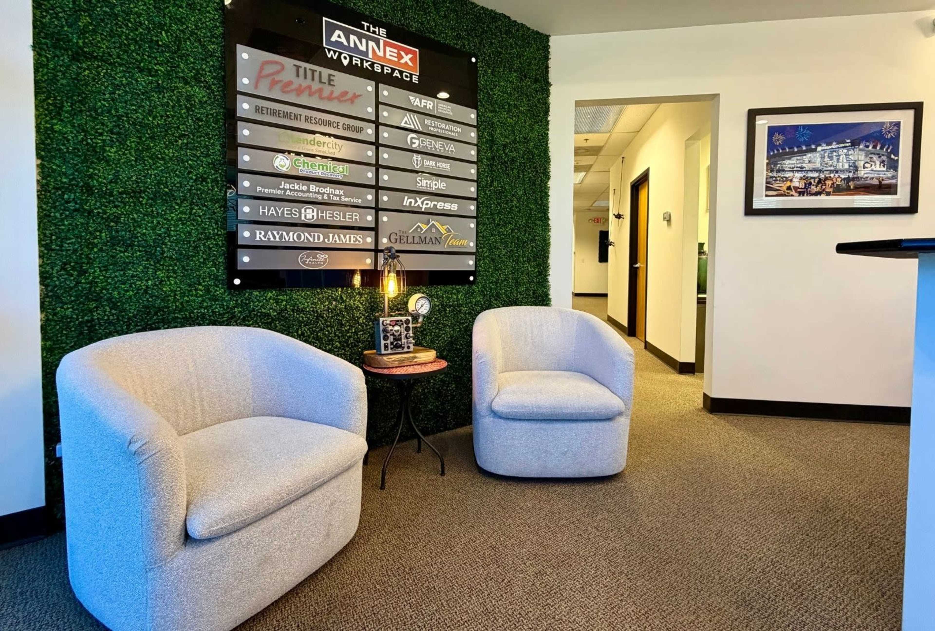 The image shows a reception area featuring two gray chairs in front of a green moss wall, with a sign listing various businesses.