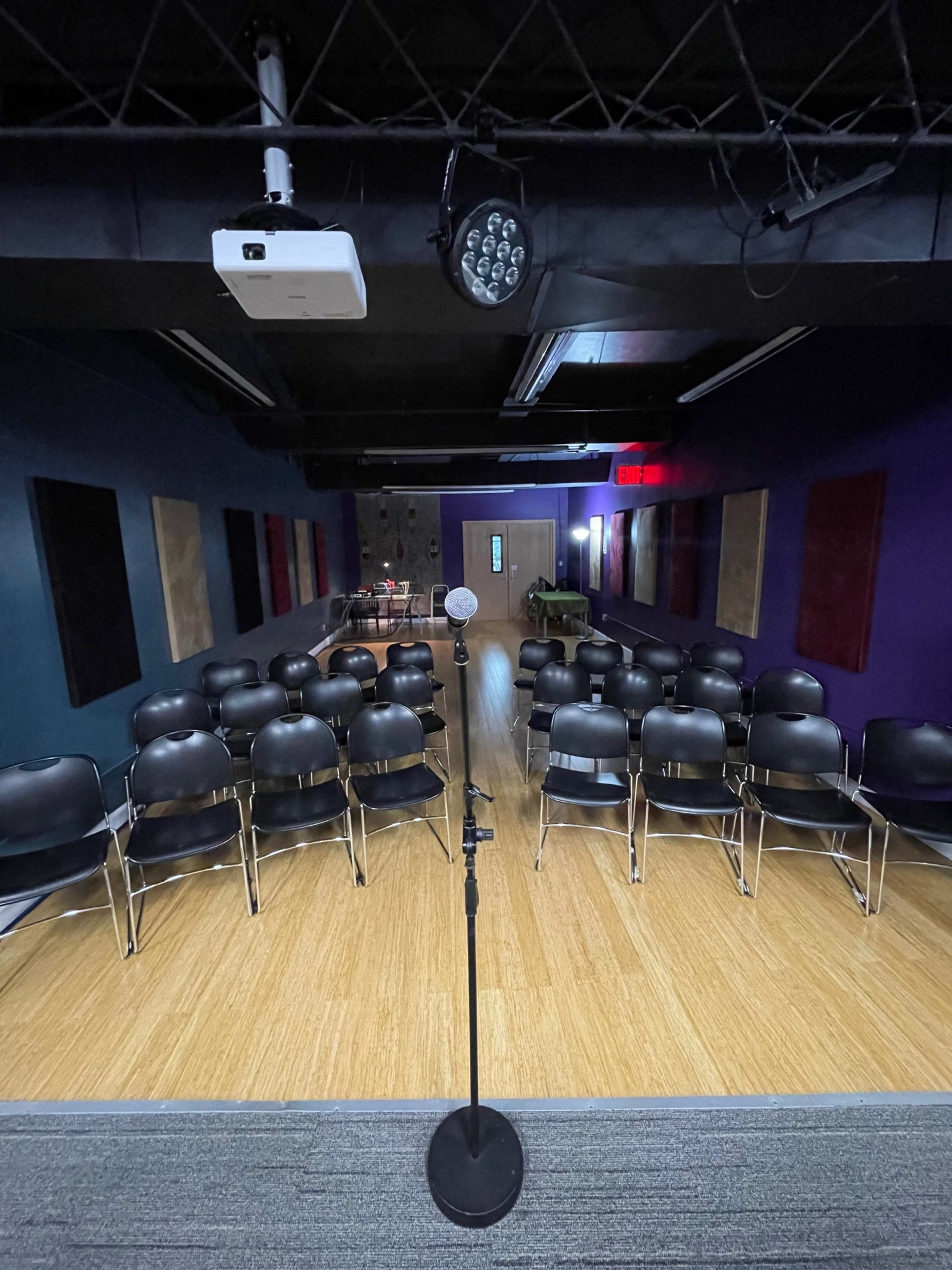 A microphone on a stand is positioned in front of a row of black chairs arranged in a performance space with purple walls and wooden flooring.