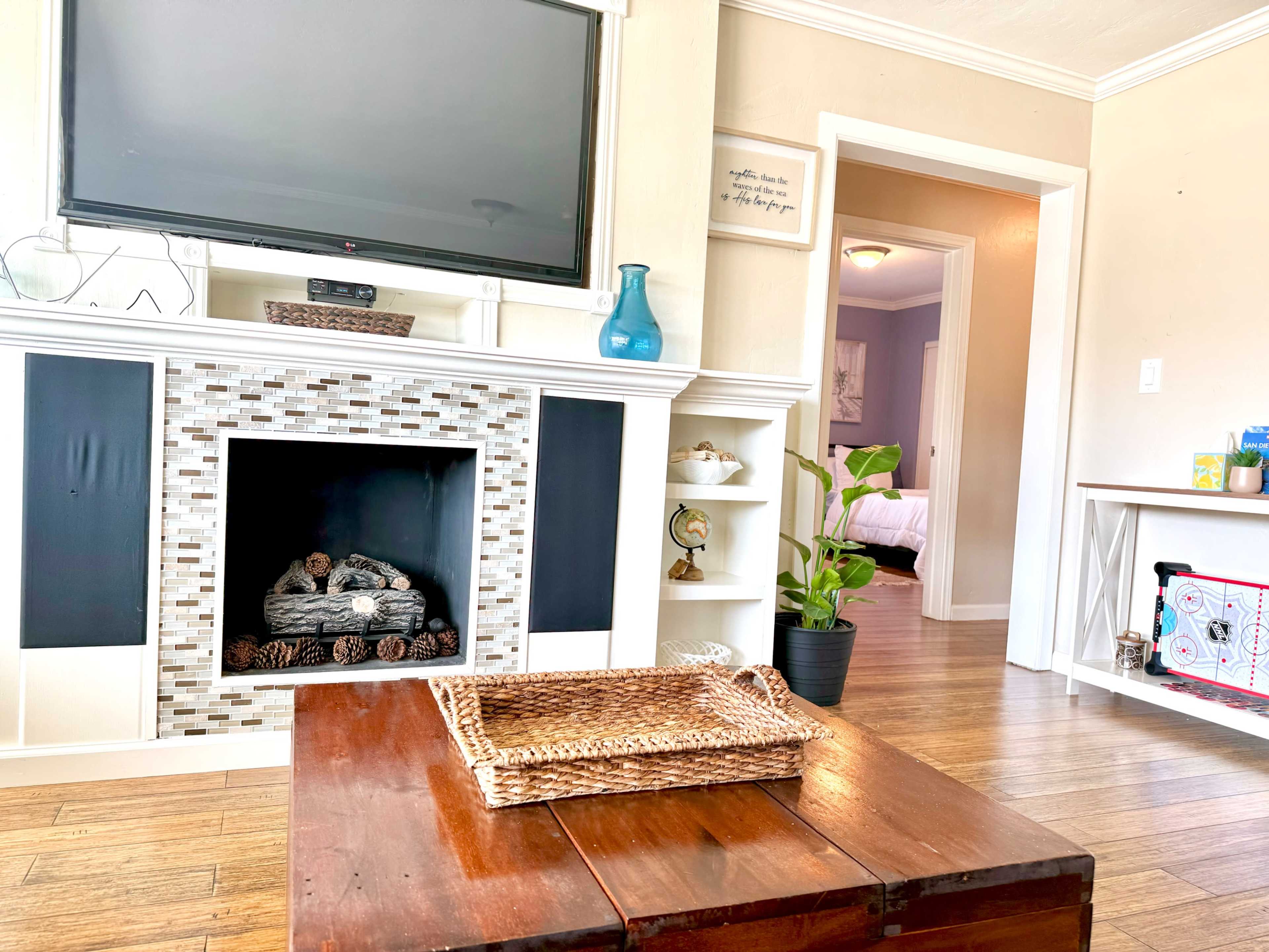 The scene features a living room with a television mounted above a mosaic-tiled fireplace, a wooden coffee table, and an entrance to another room visible in the background.