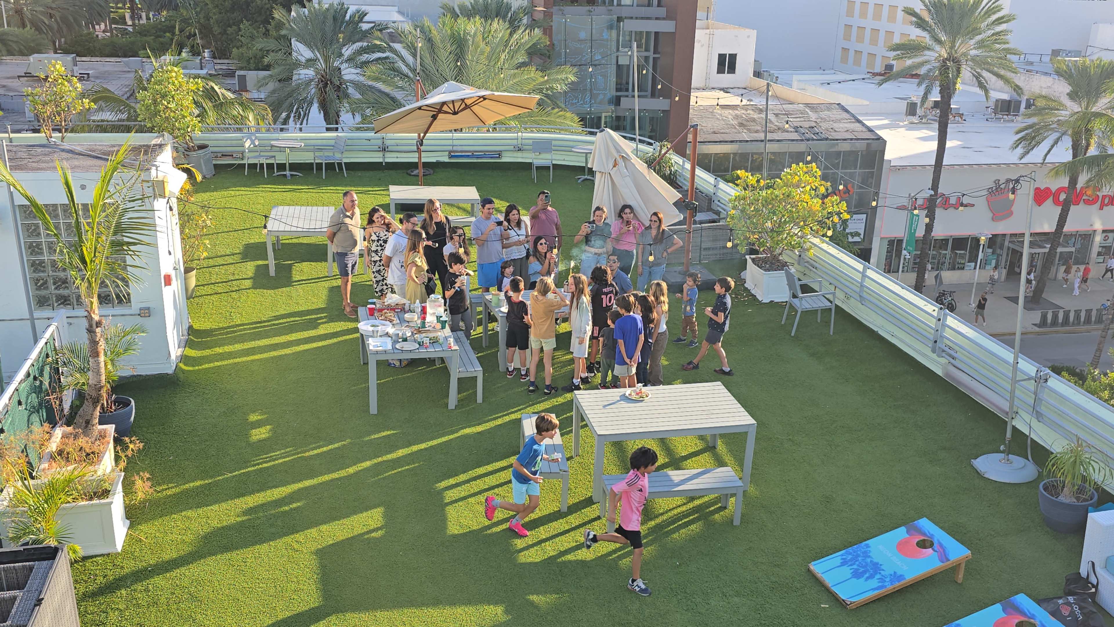 A group of people is gathered on a rooftop, enjoying a social event with tables set up for food and games in a lush, green environment.