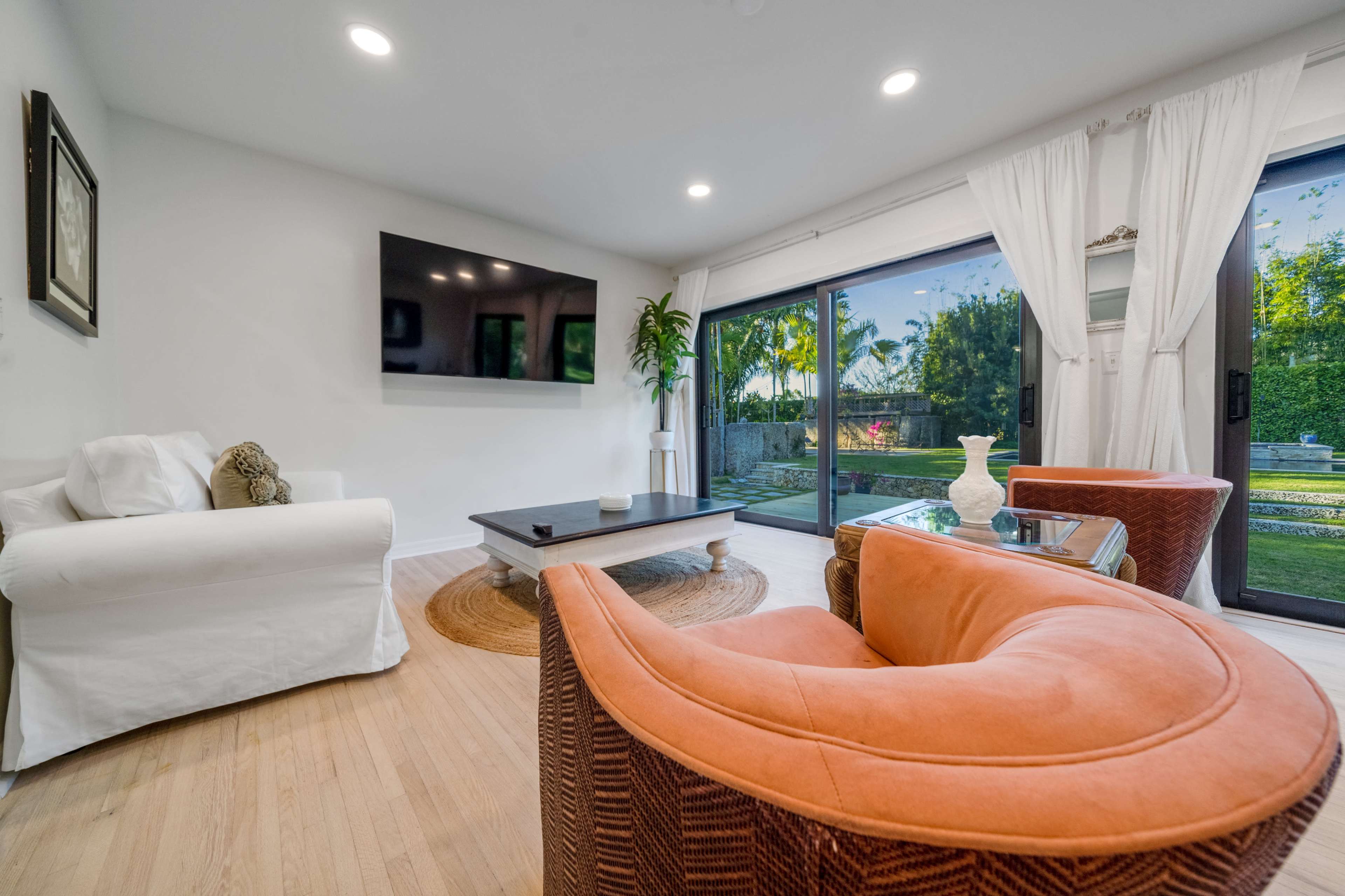 A living room features a white sofa, an orange armchair, a dark coffee table, and large sliding glass doors that open to a landscaped outdoor area.