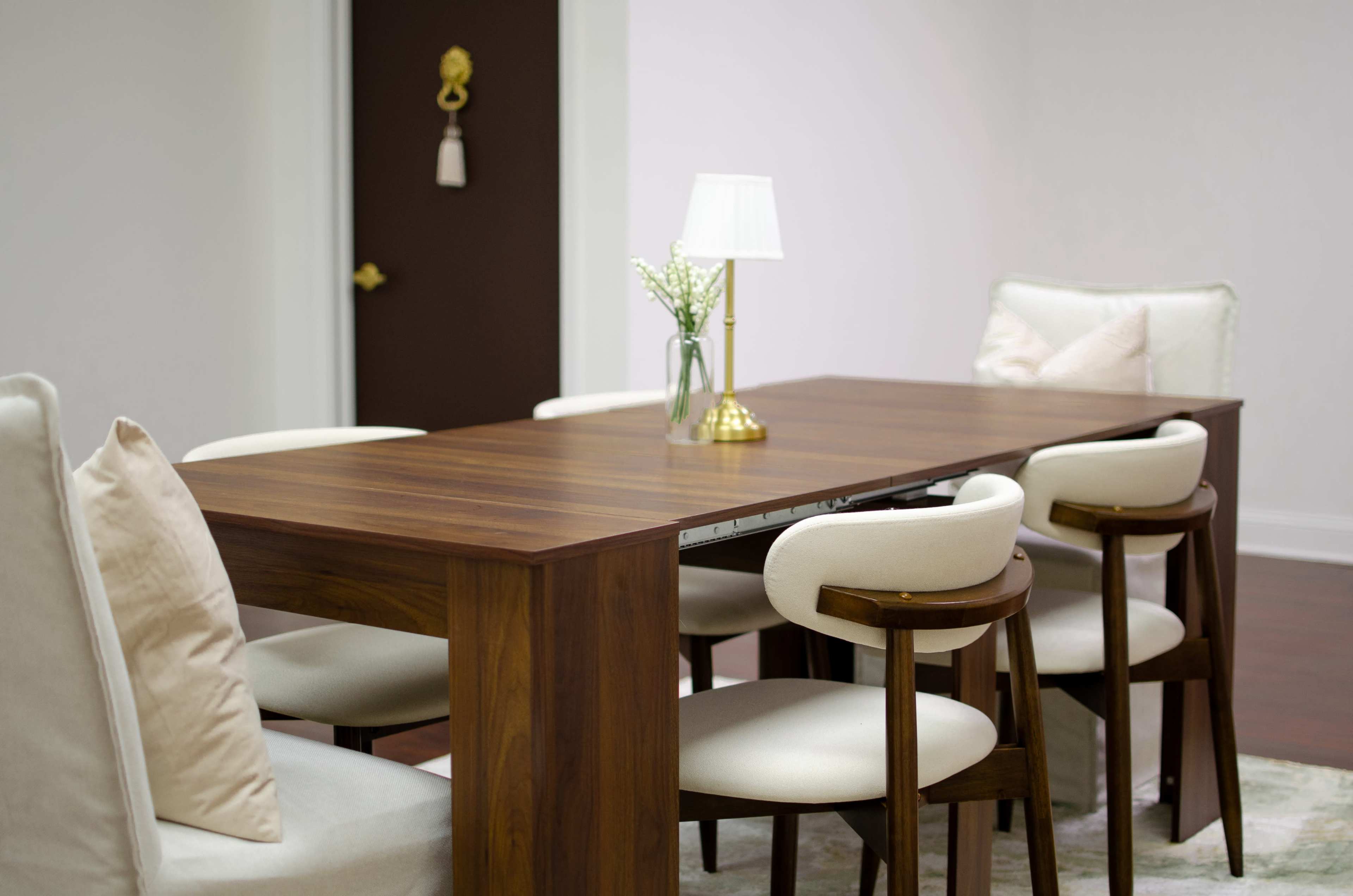 A wooden dining table is set with six cream-colored chairs in a minimalist room featuring soft lighting and a small vase with flowers on the table.