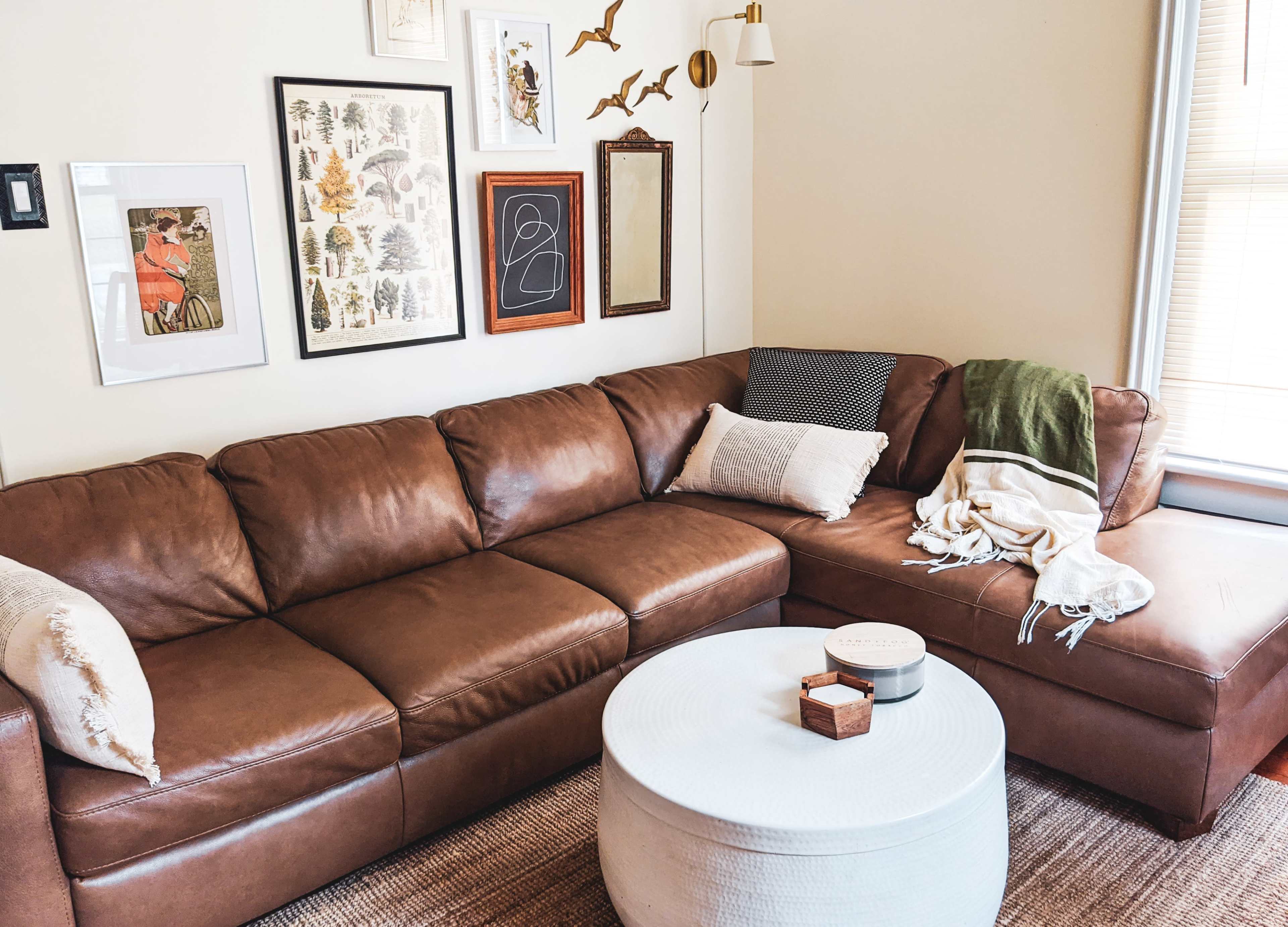 The image features a brown leather sectional sofa positioned against a wall adorned with framed artwork, alongside a round white coffee table and various decorative items.
