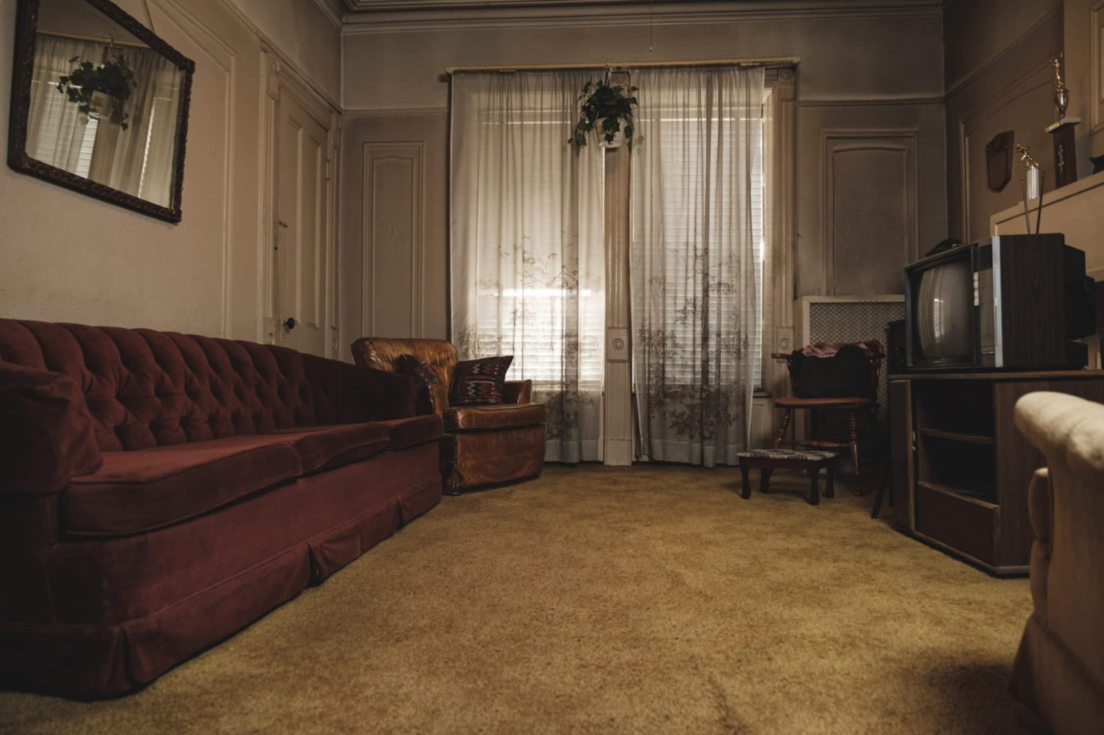 The scene shows a vintage living room with a red velvet sofa, an armchair, a television, and large curtains partially covering the windows.