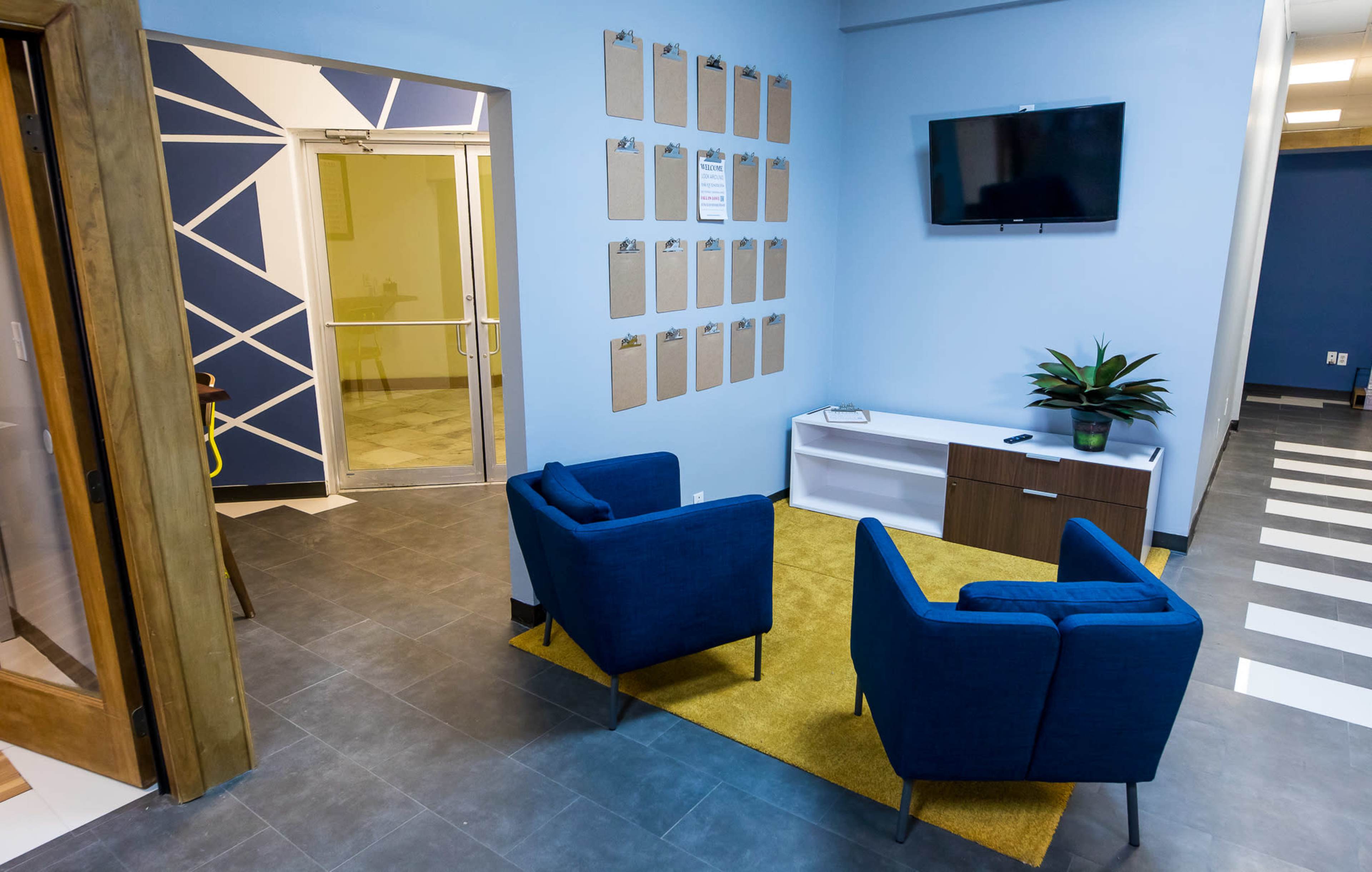 A modern waiting area with two blue armchairs, a white cabinet, a wall-mounted television, and a number of clipboards arranged on the wall.