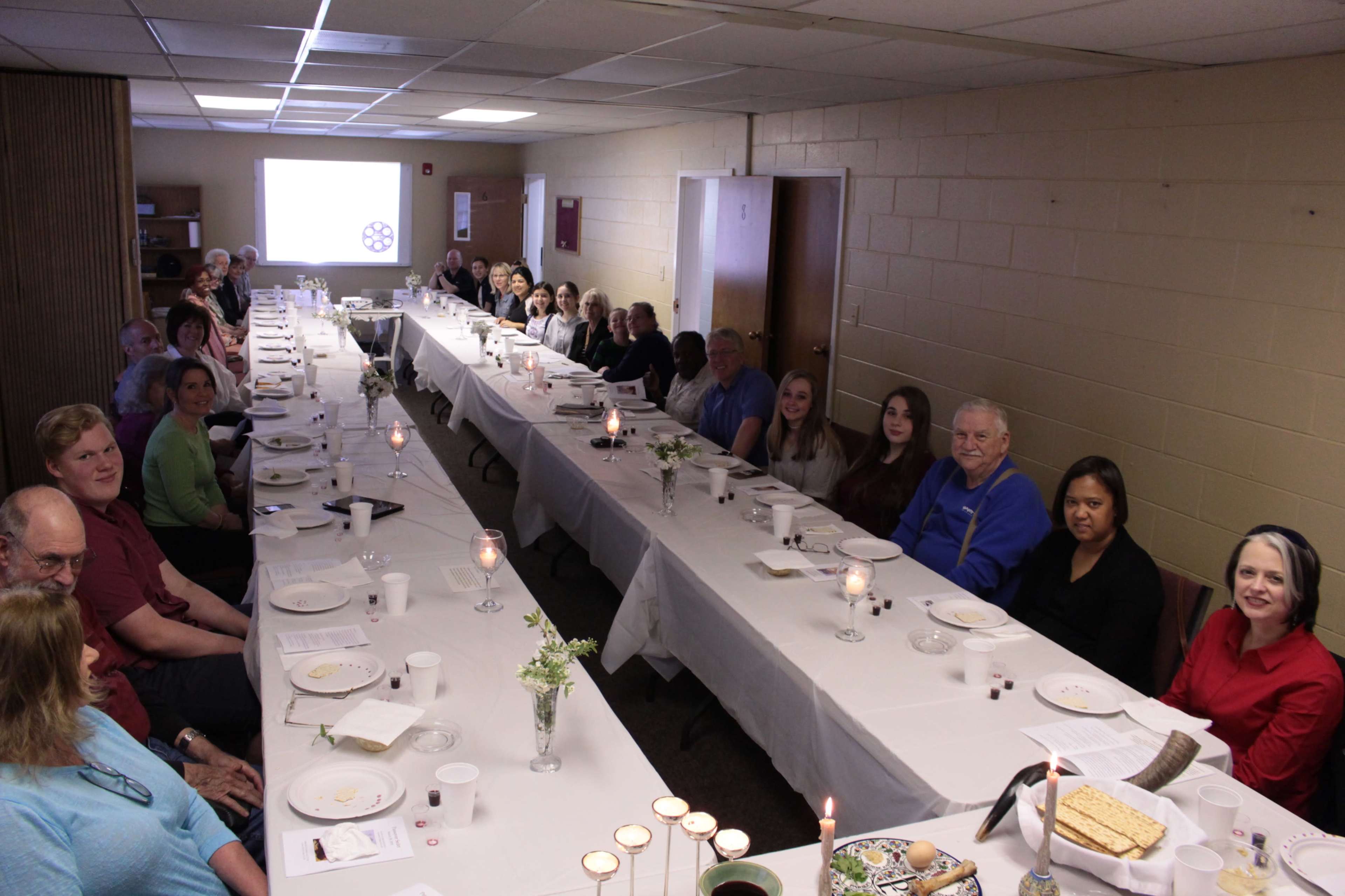A long table is set for a gathering, with people seated on both sides, in a well-lit room featuring decorative candles and plates.
