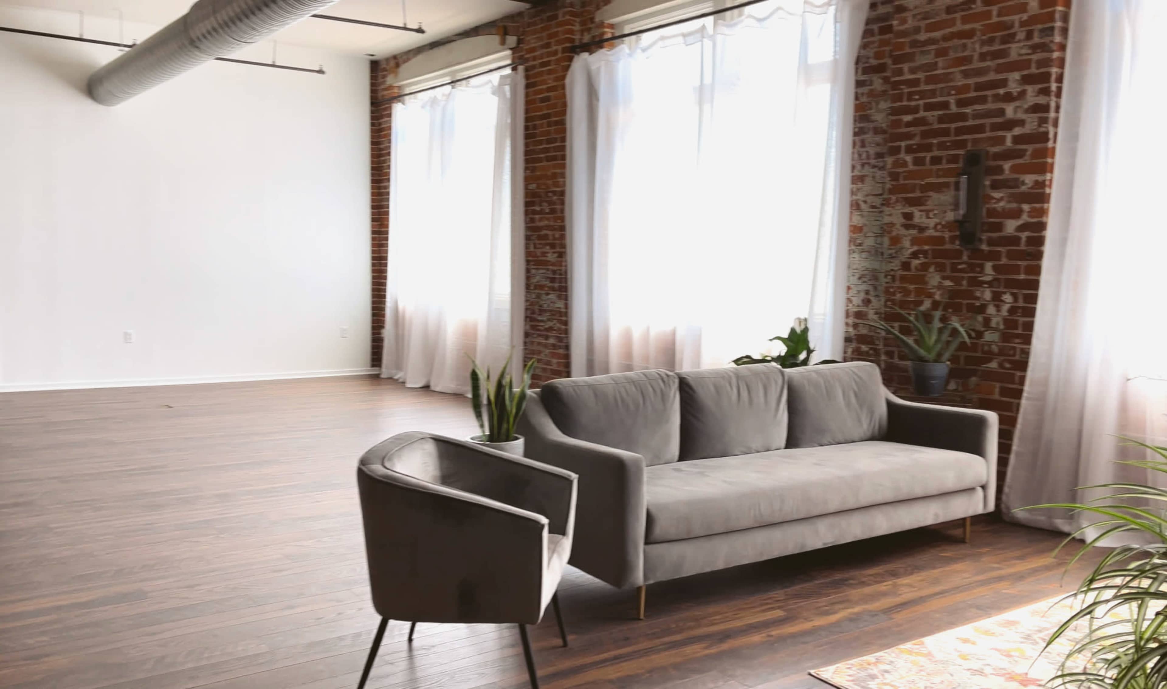 The image shows a spacious room with large windows, exposed brick walls, a gray sofa, and a chair, featuring wooden floors and potted plants.