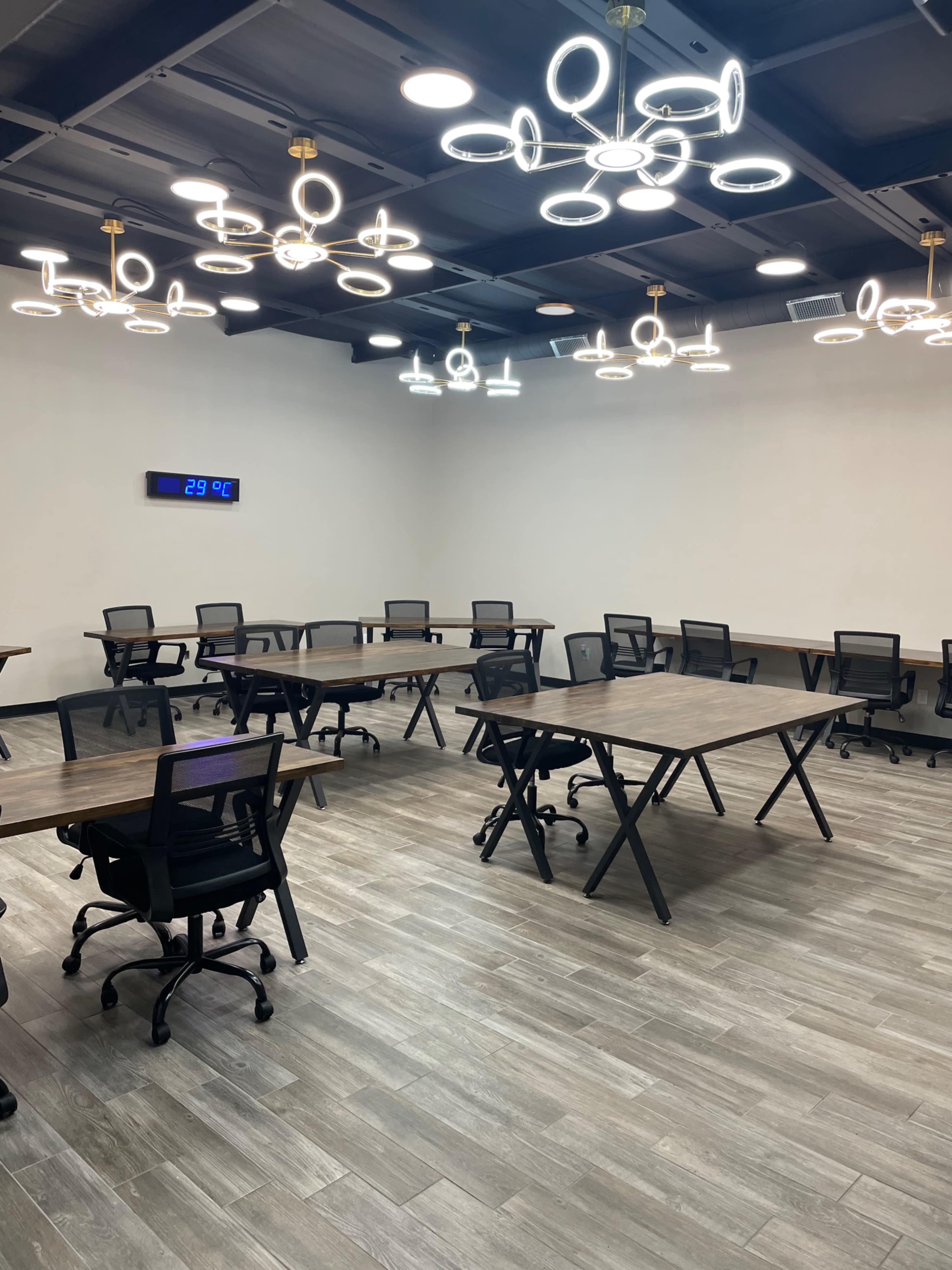 A spacious, empty room features several tables and chairs arranged for meetings, with modern circular light fixtures and a digital temperature display on the wall.