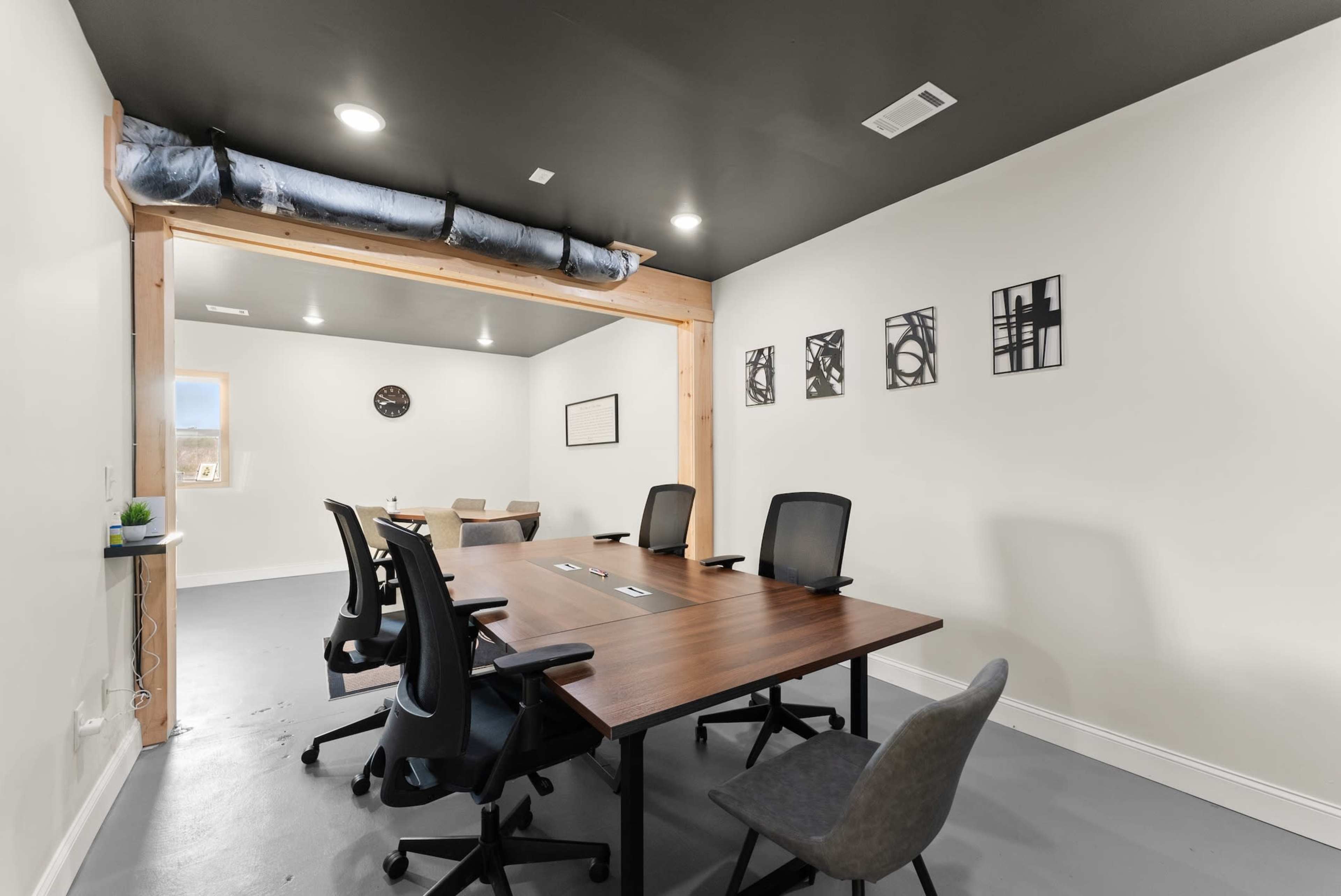 A conference room features a large wooden table surrounded by office chairs, with a clock and framed art on the walls.