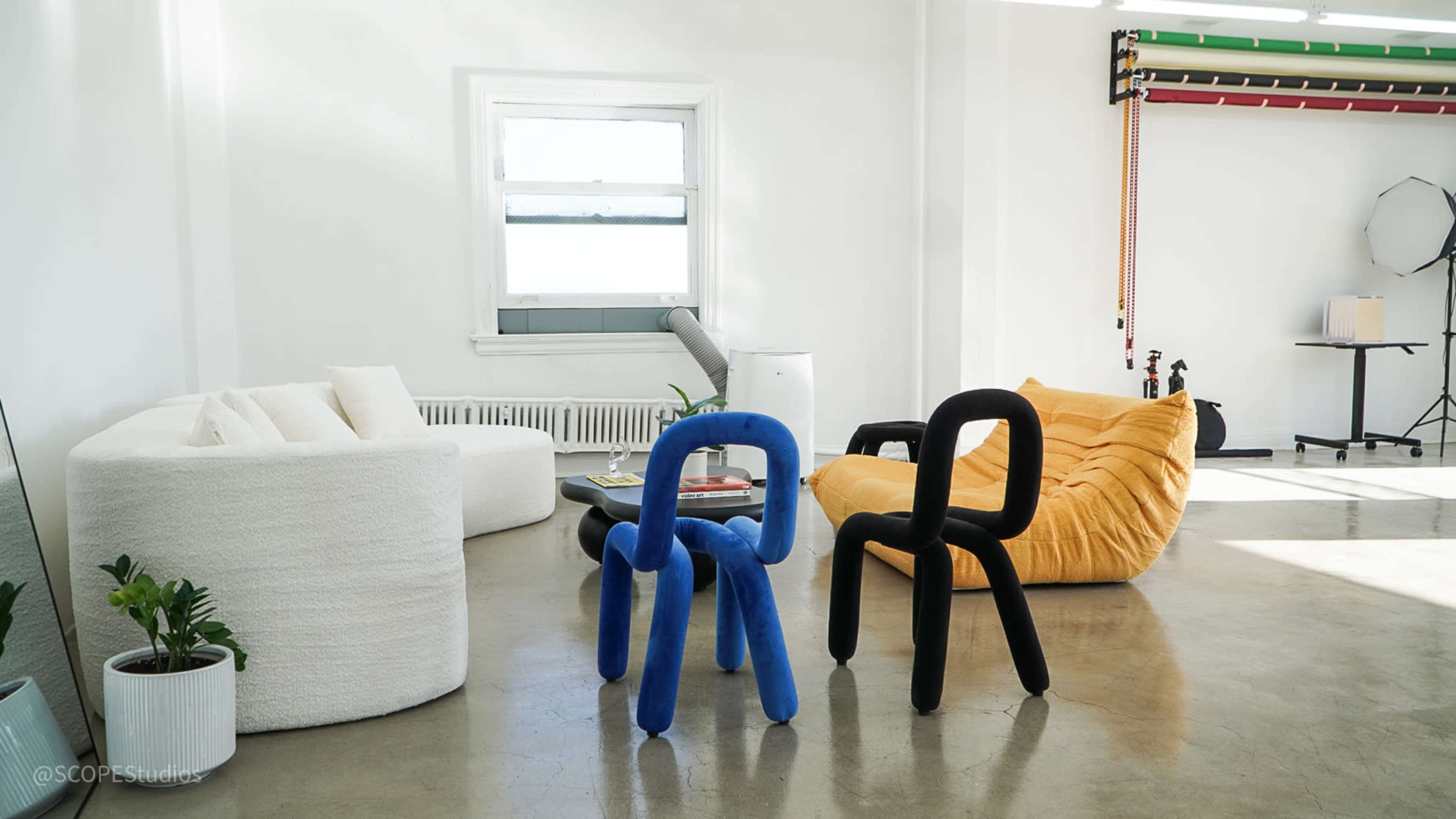 The image features a spacious, well-lit room with modern furniture, including a white circular sofa, a yellow bean bag chair, and colorful sculptural chairs in black and blue.