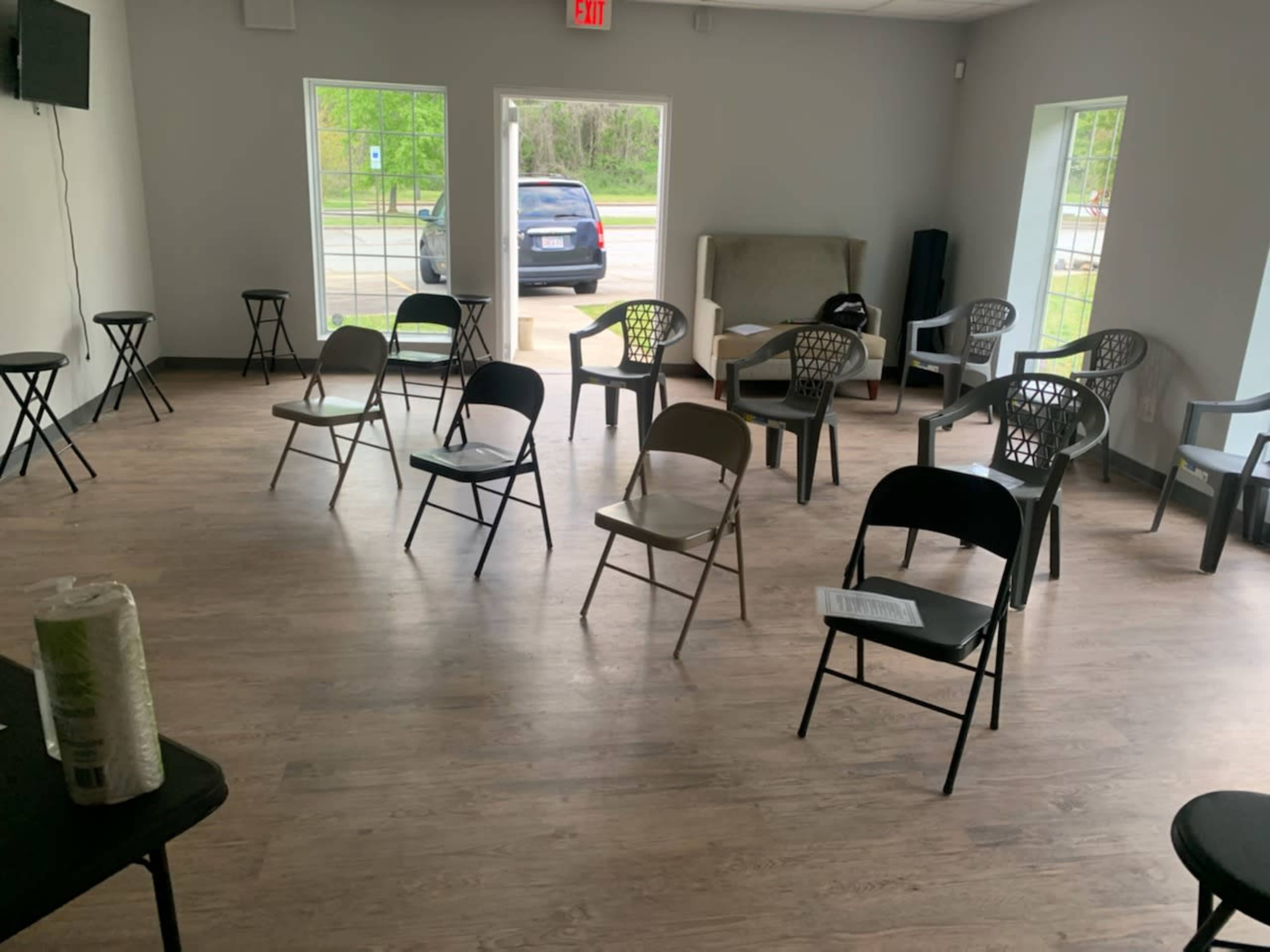 A room with wooden flooring contains several chairs arranged in a scattered manner, with a few small tables and a couch against the wall.