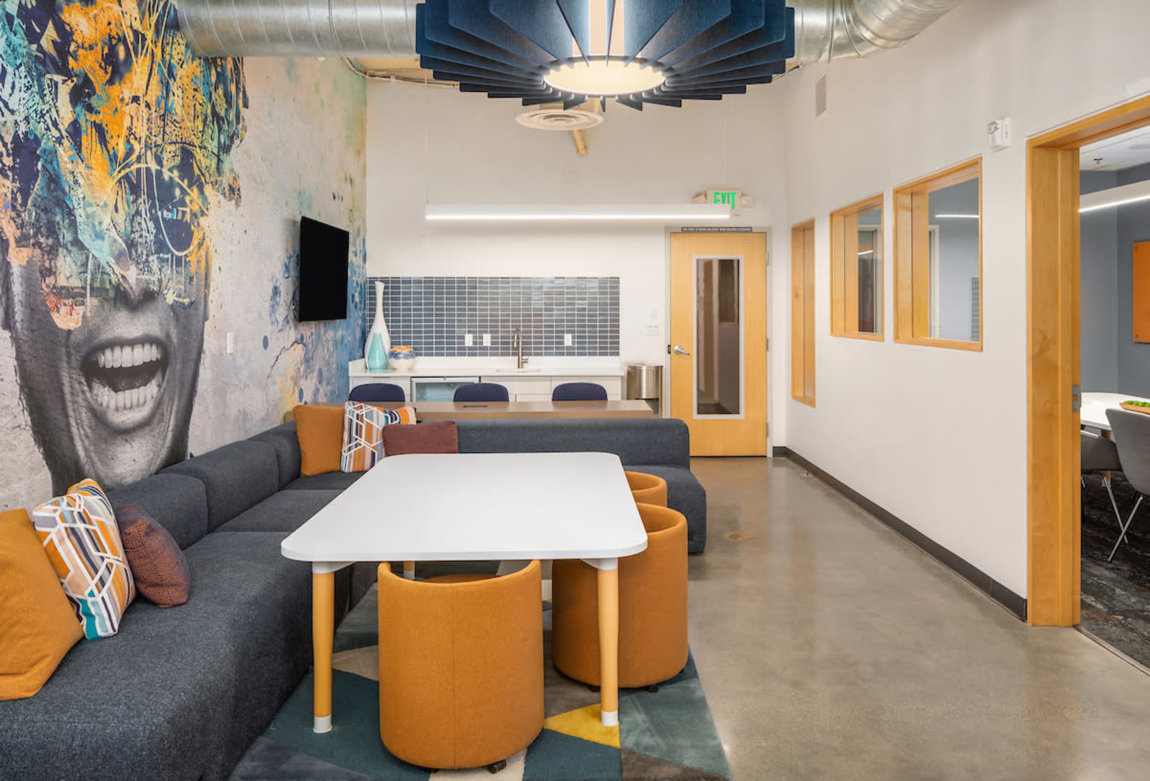 A modern communal space with a gray couch, a white table surrounded by colorful chairs, and a vibrant mural on the wall.