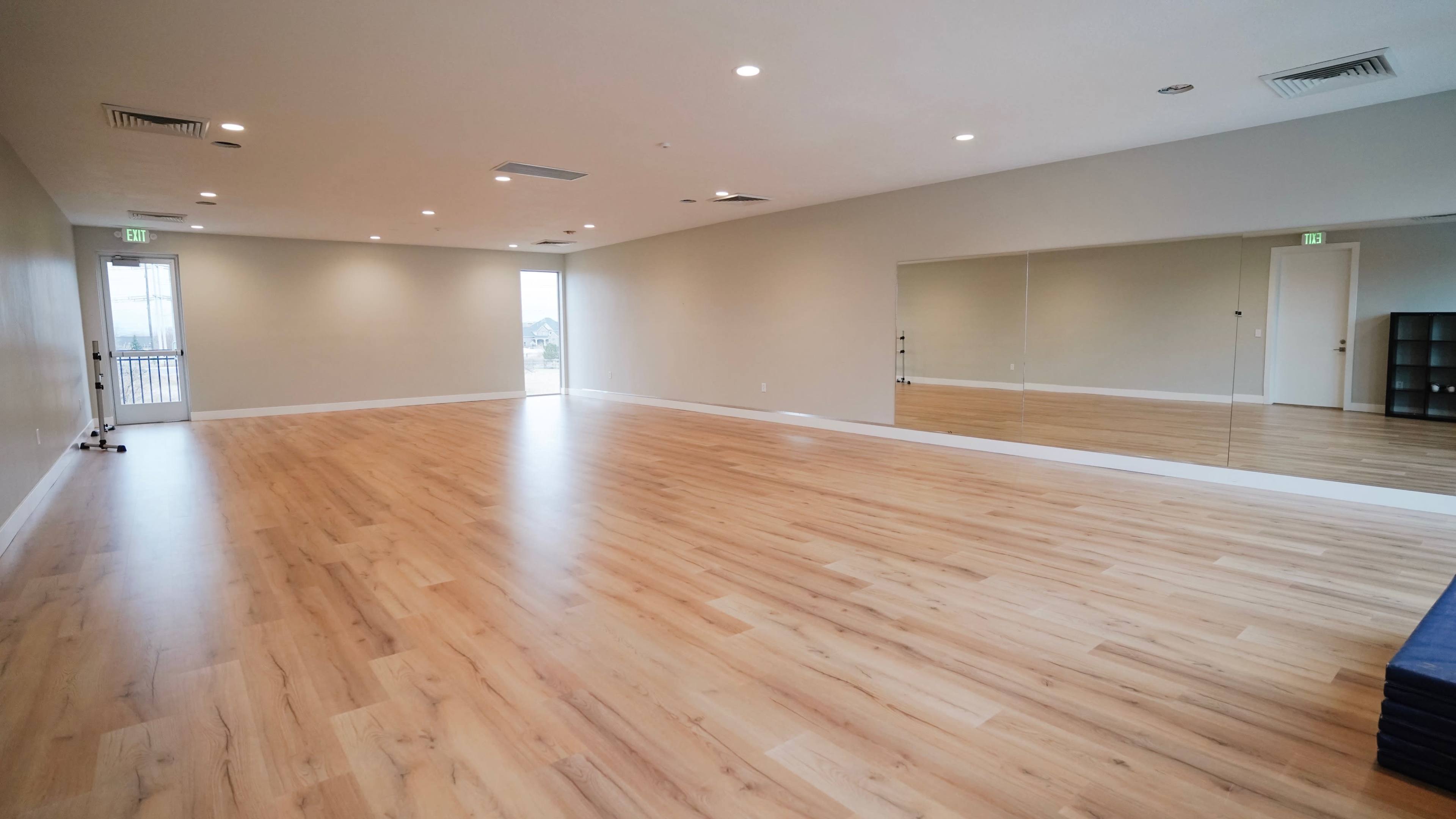 A spacious, empty room with wooden floors, mirrors on one wall, and natural light coming through large windows.