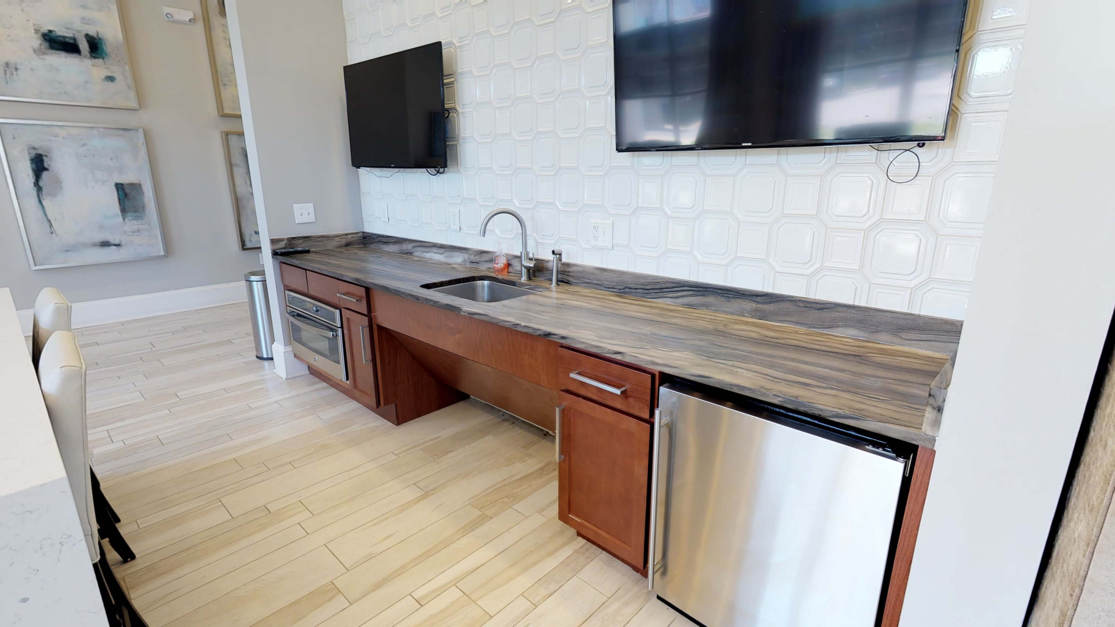 A modern kitchen area with a wooden countertop, a sink, an oven, and a stainless steel refrigerator, alongside two mounted flat-screen TVs.