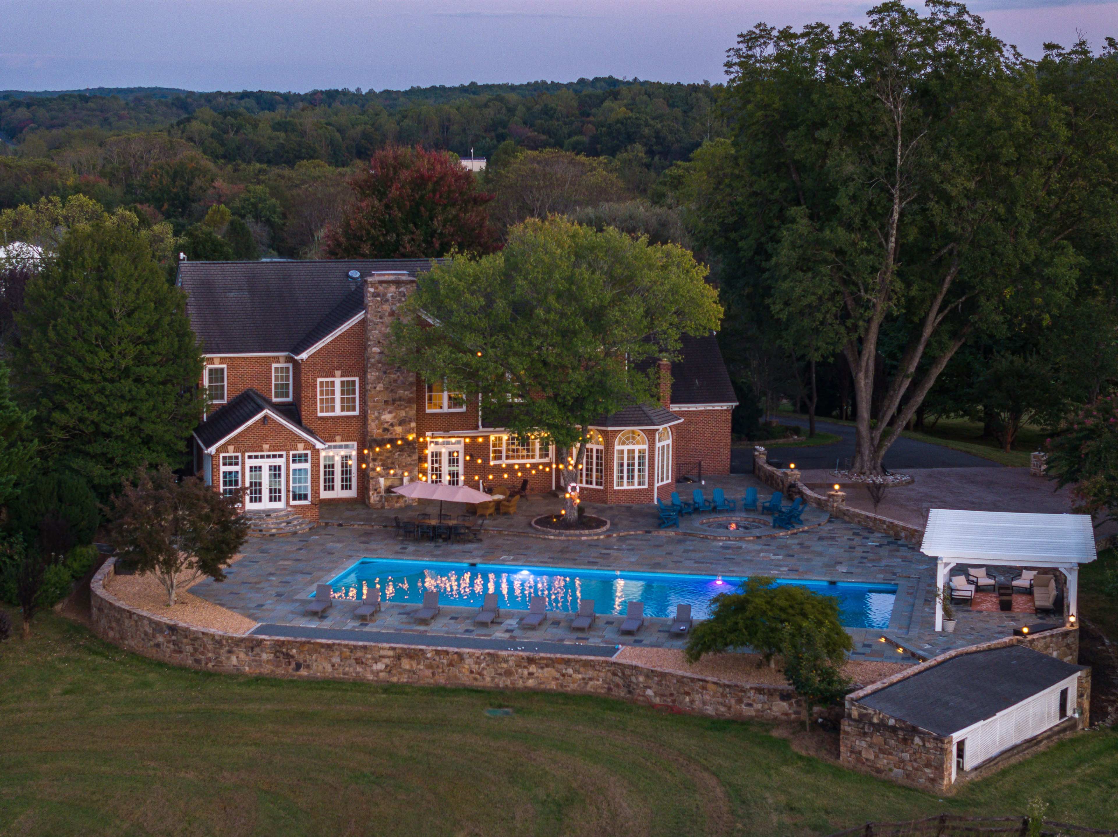 The image shows a large brick house with a swimming pool and patio area surrounded by trees and landscaping, captured from an aerial perspective during twilight.