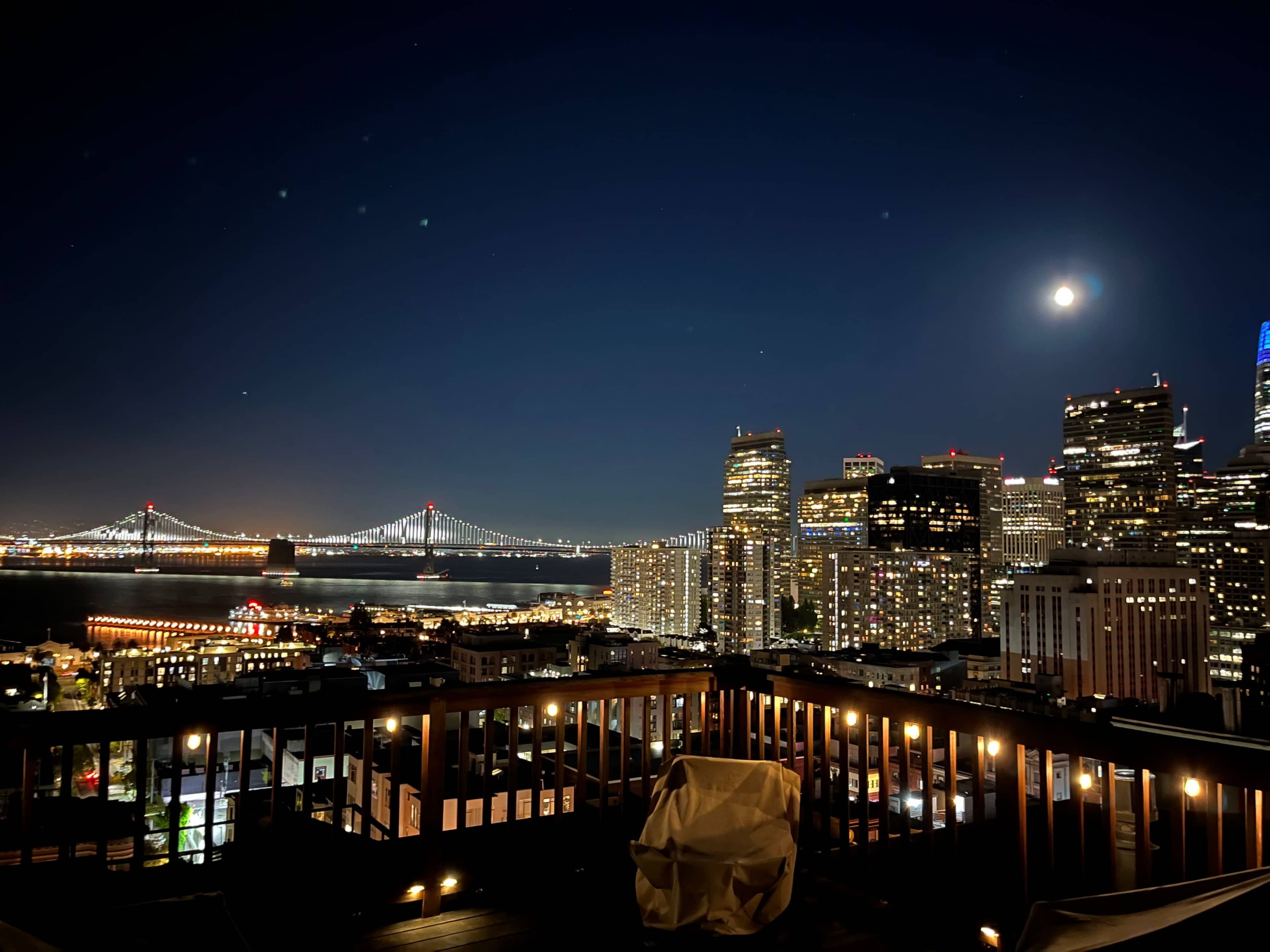 A night view of a city skyline with a bridge illuminated in the background and a full moon above.
