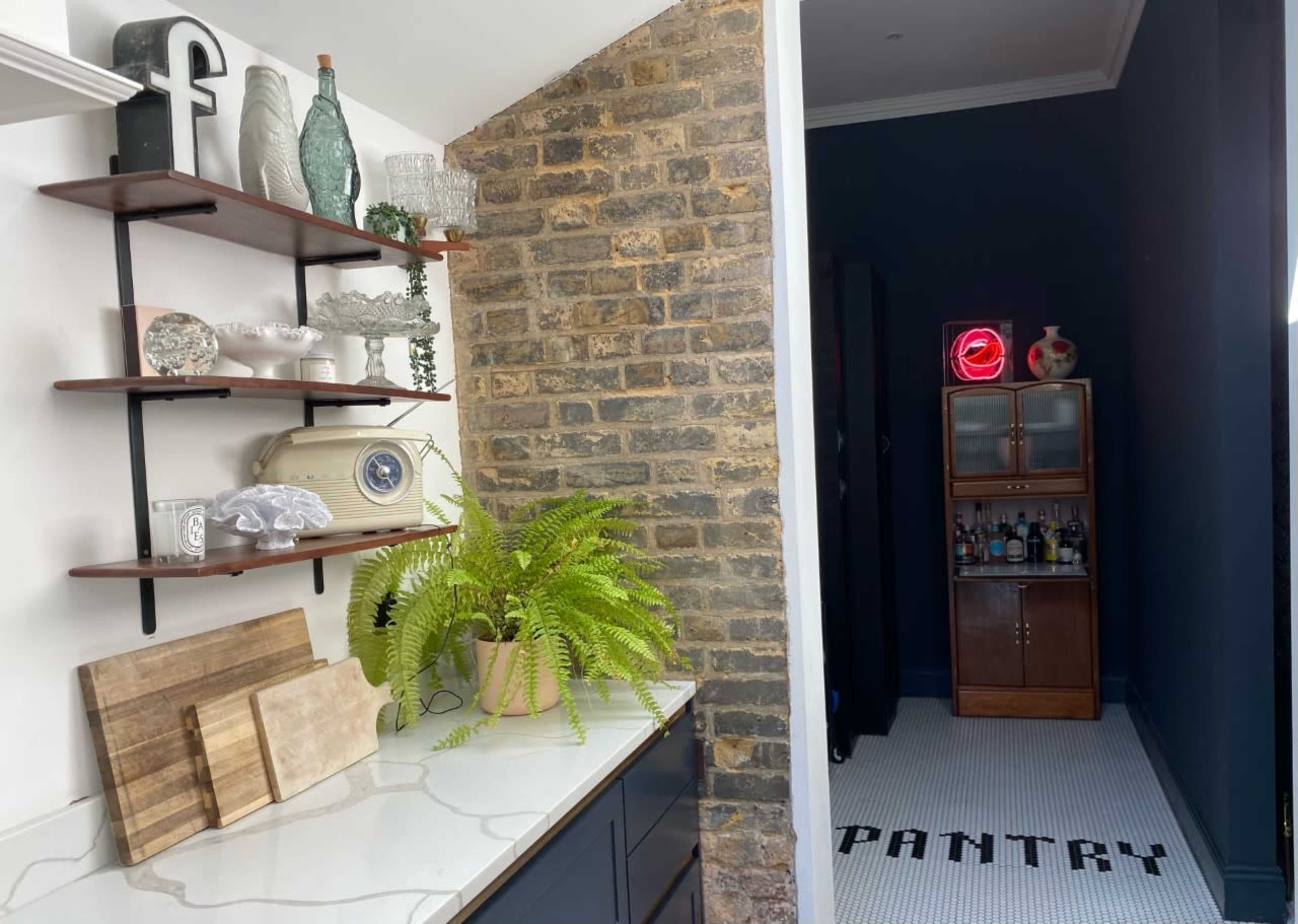 The image shows a kitchen area with wooden shelves displaying glassware and a vintage clock, leading to a pantry with a neon sign.
