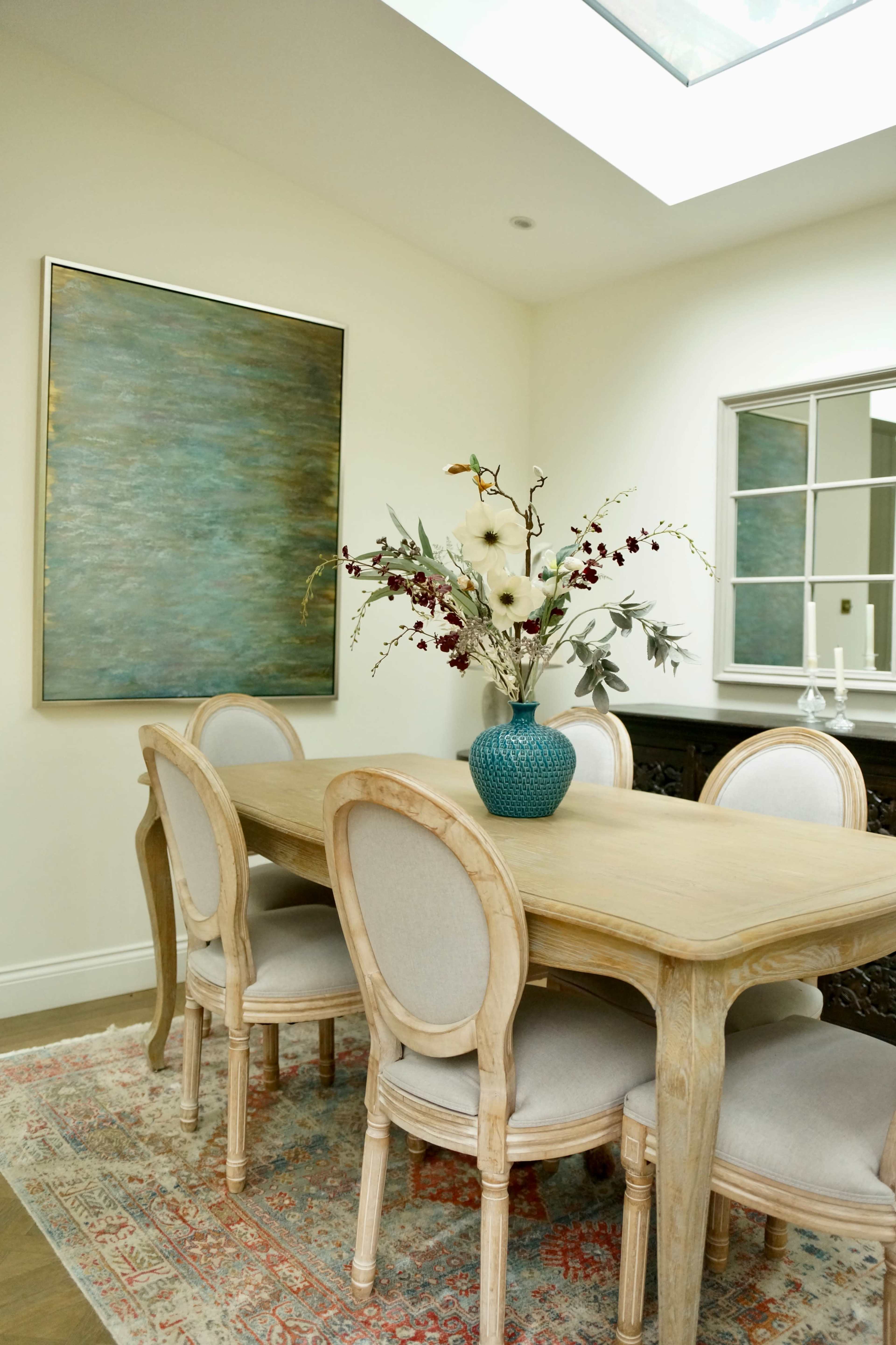 A wooden dining table with six upholstered chairs is centered in a well-lit room featuring a large piece of abstract art on the wall and a vase of flowers on the table.