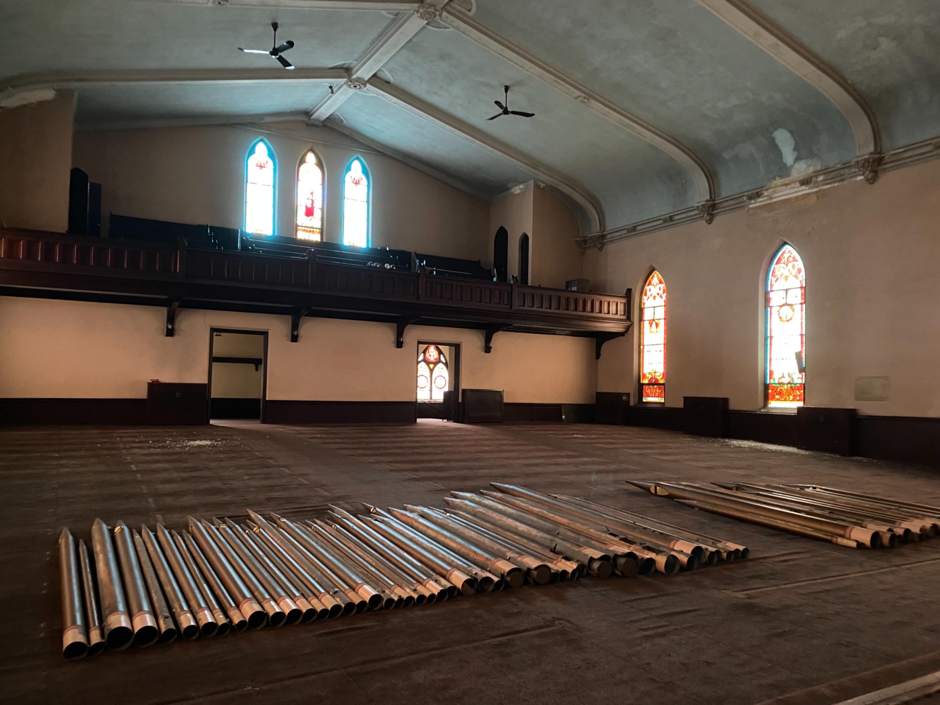 A spacious, empty hall with stained glass windows and metal pipes arranged neatly on the floor.