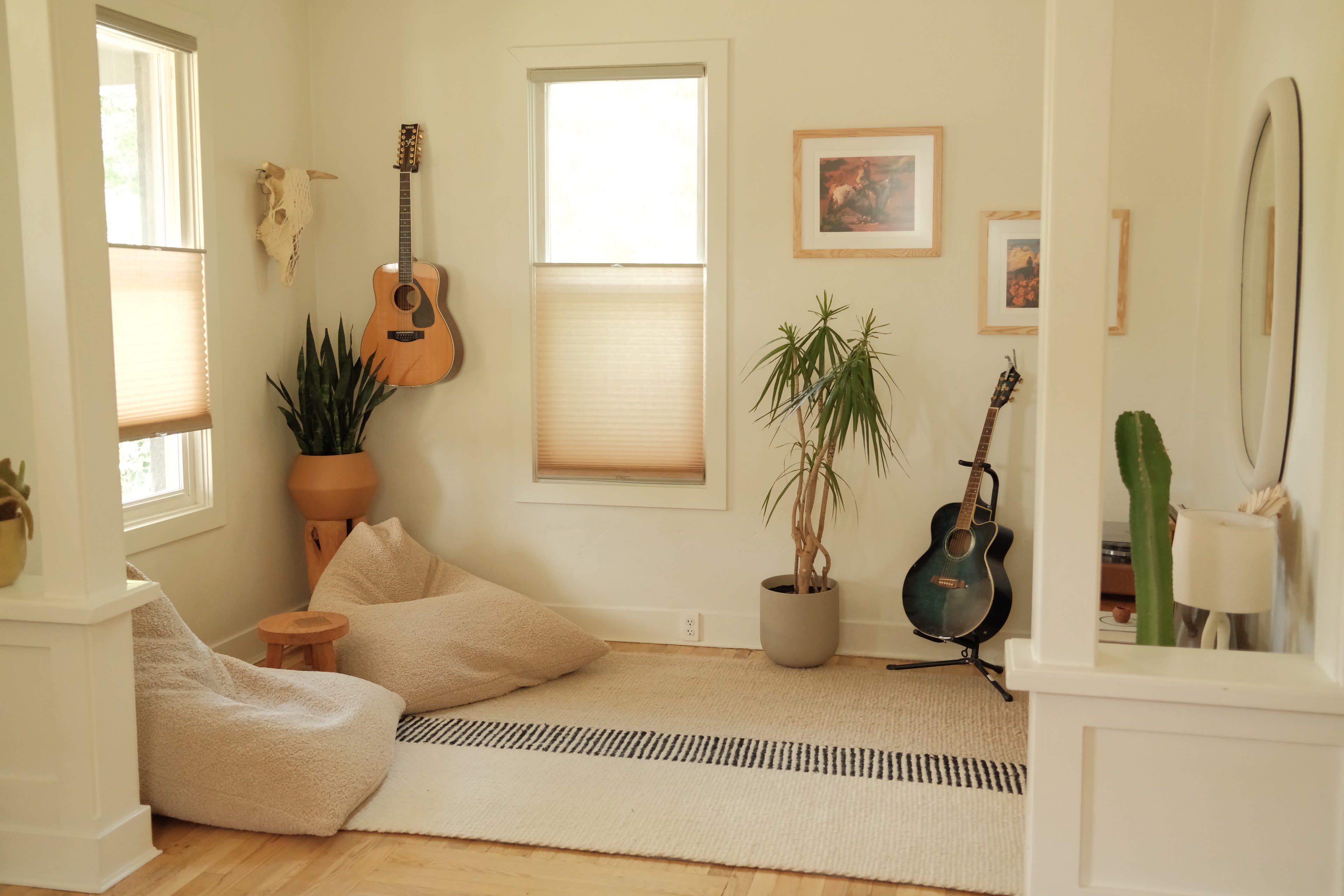 The image shows a cozy corner of a room featuring two guitars, a plant, bean bag seating, and a textured rug.