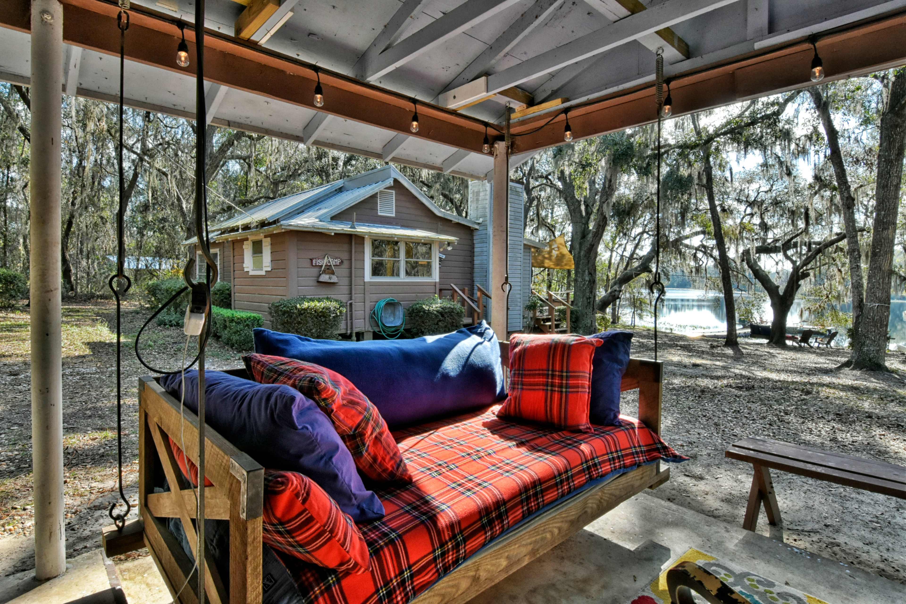 A swing seating area with colorful cushions overlooks a lake and a house surrounded by trees.