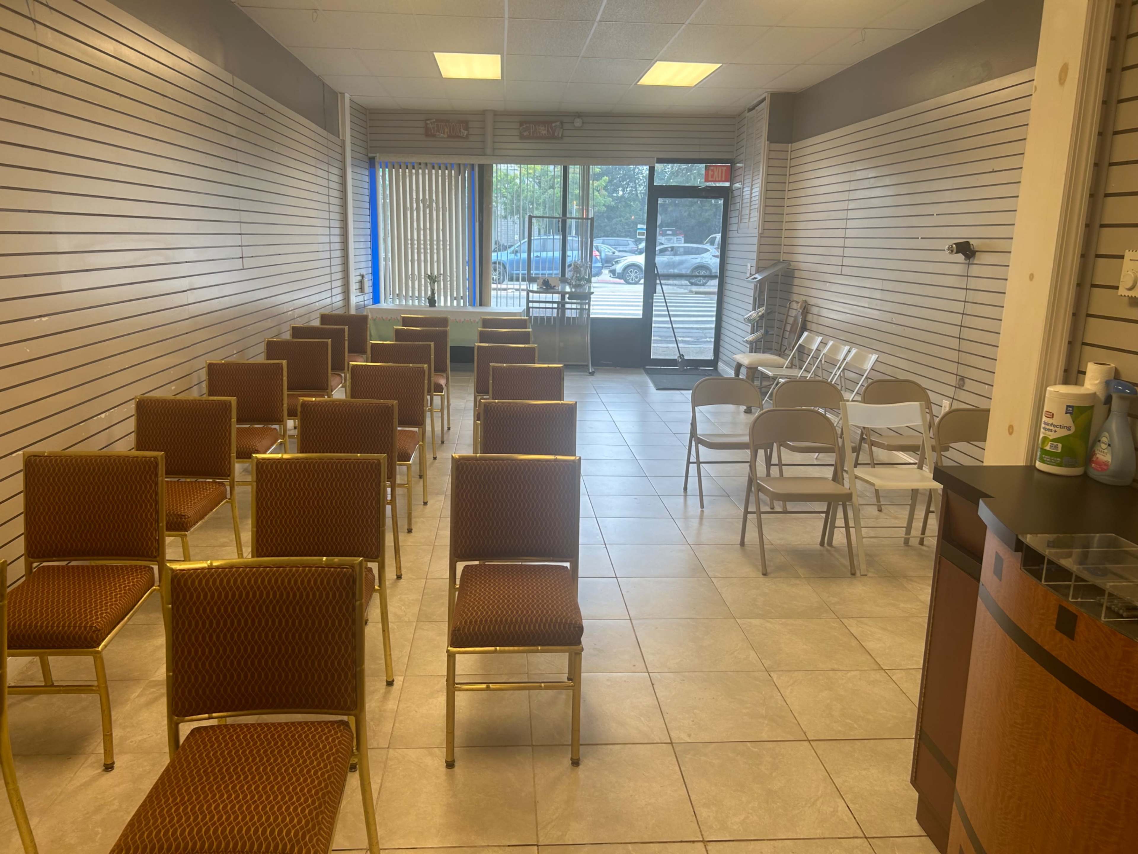 An empty room with rows of brown upholstered chairs and white folding chairs arranged for an event, with large windows providing natural light.