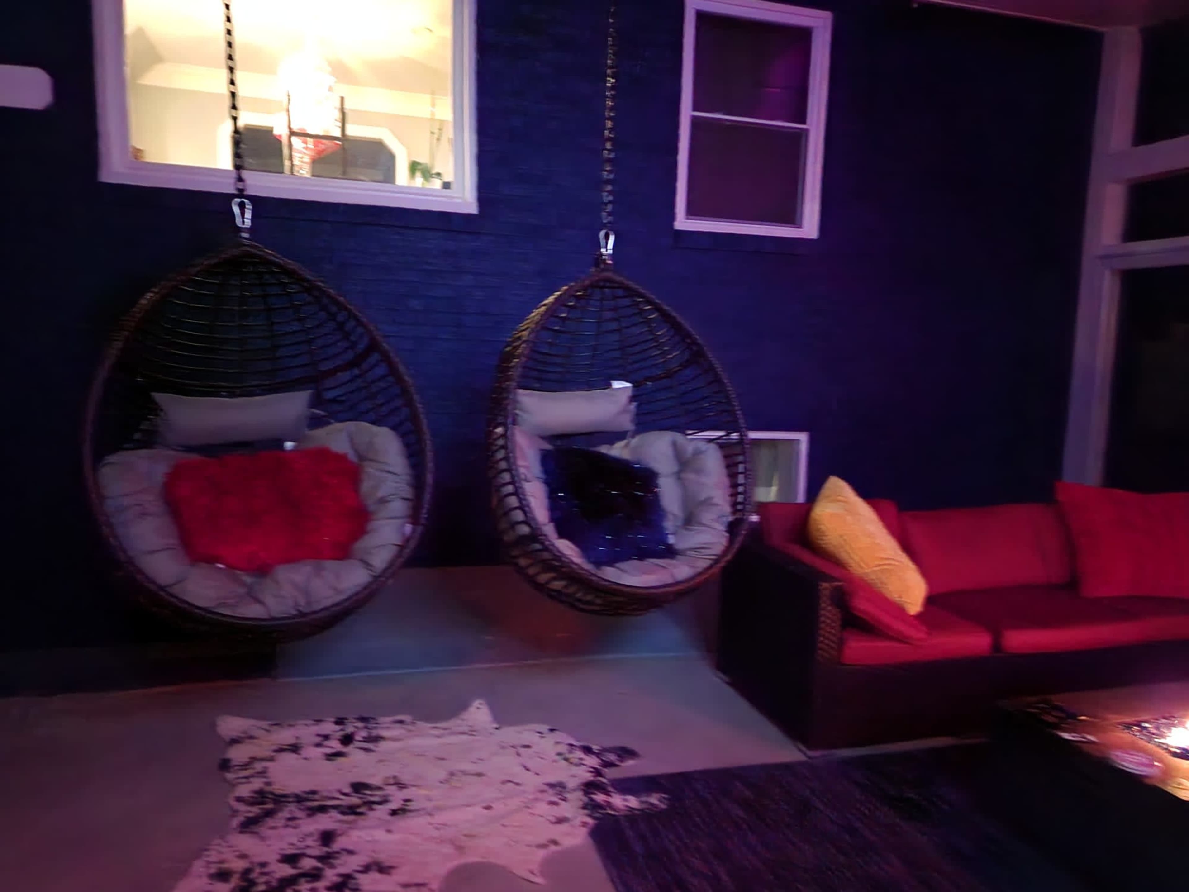 Two hanging basket chairs and a red sofa arranged on a patio with a dark blue wall and a visible window.