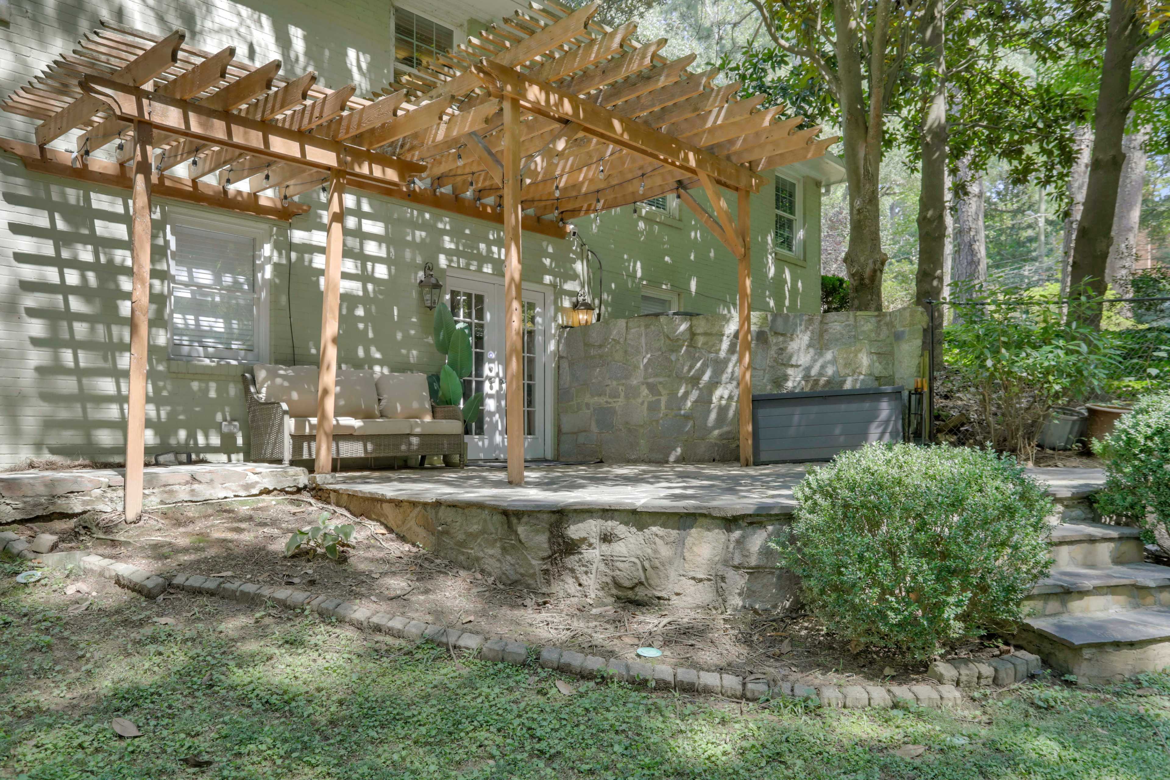 A patio area with a wooden pergola, a stone wall, and neatly trimmed shrubs in a residential backyard.