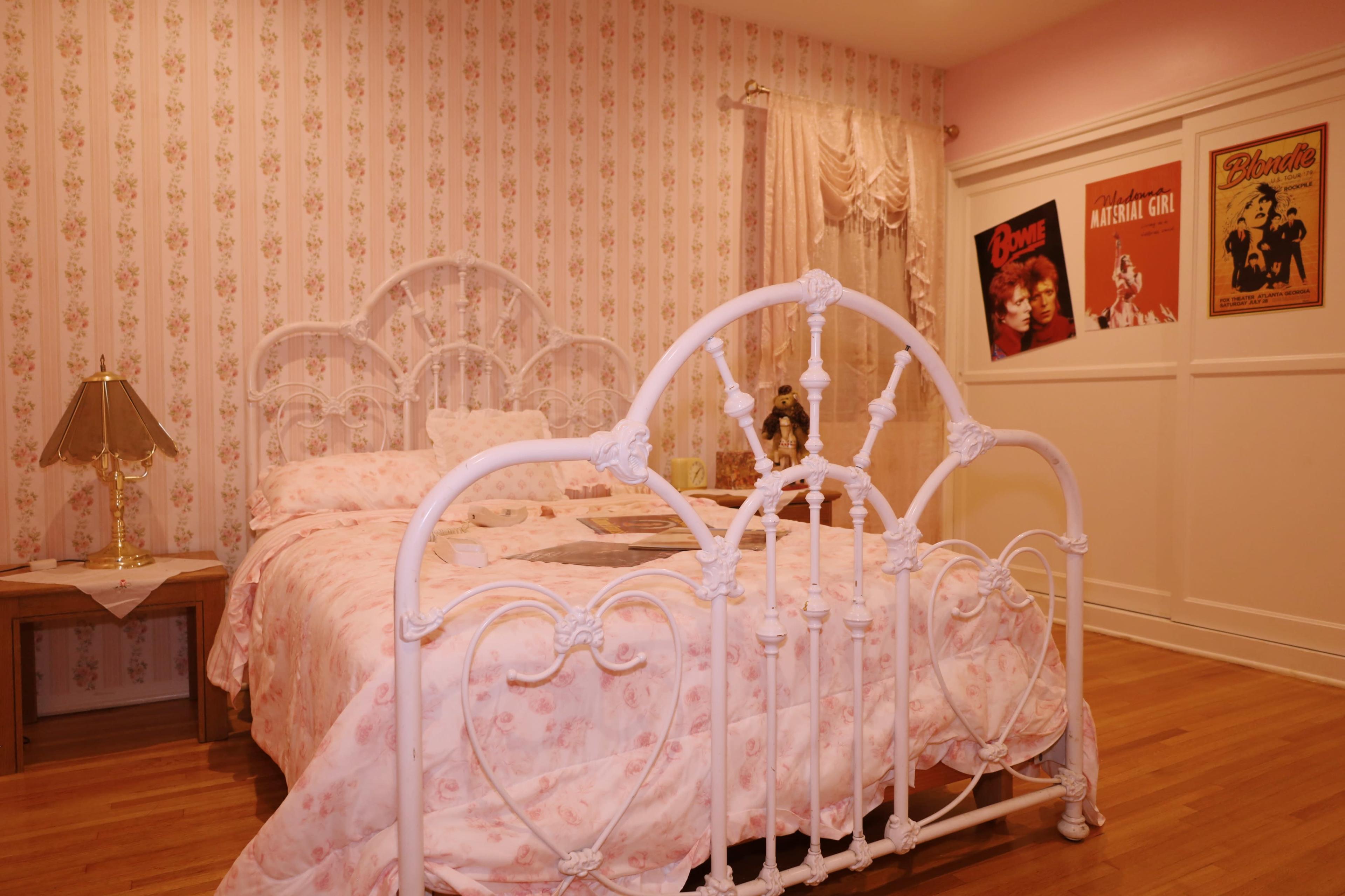 The image shows a vintage-style bedroom featuring a white wrought iron bed with pink floral bedding, a lamp on a nightstand, and floral wallpaper along with framed posters on the wall.