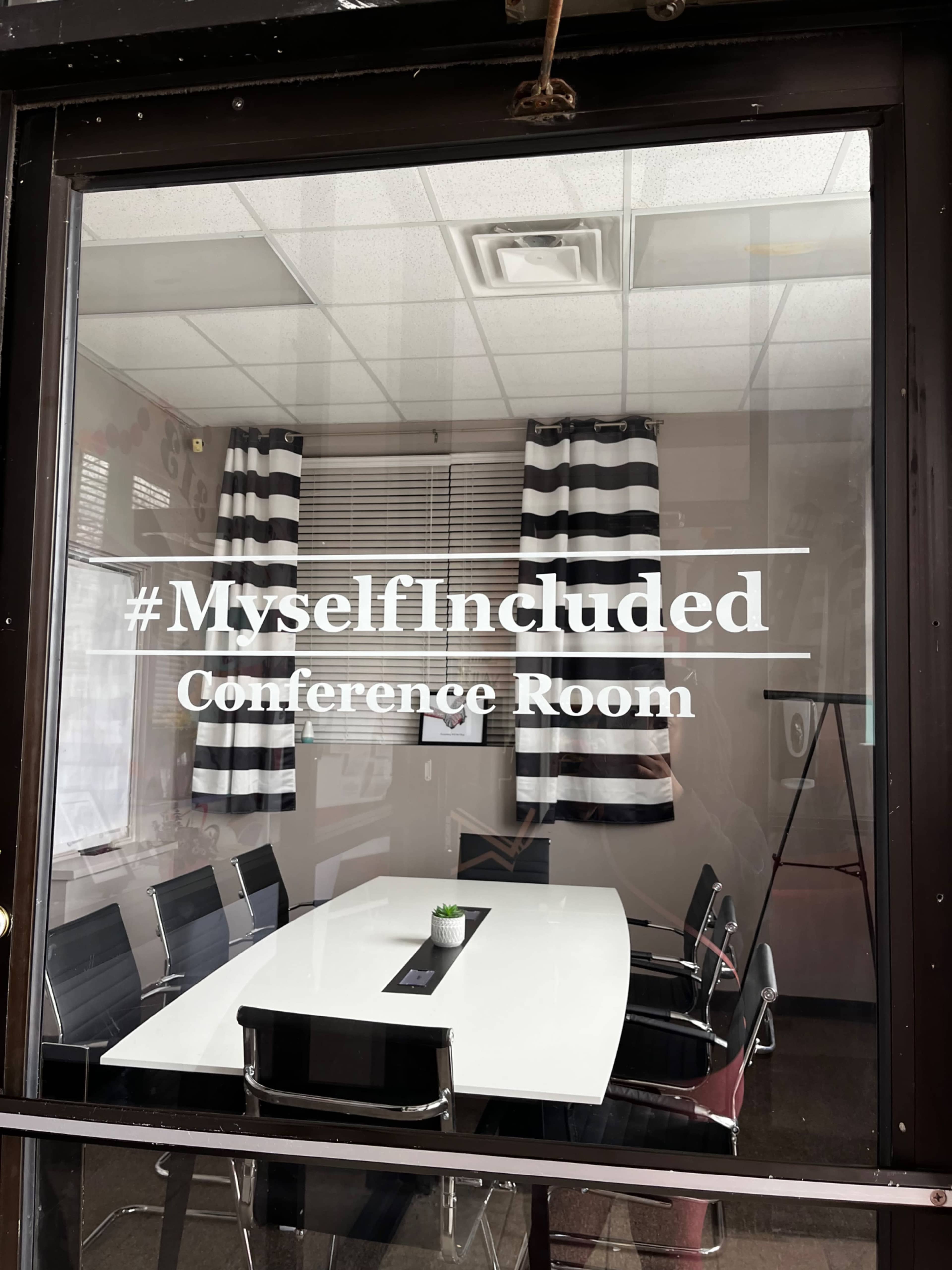 The image shows a conference room with a glass window labeled "#MyselfIncluded Conference Room," featuring a white table and black chairs.