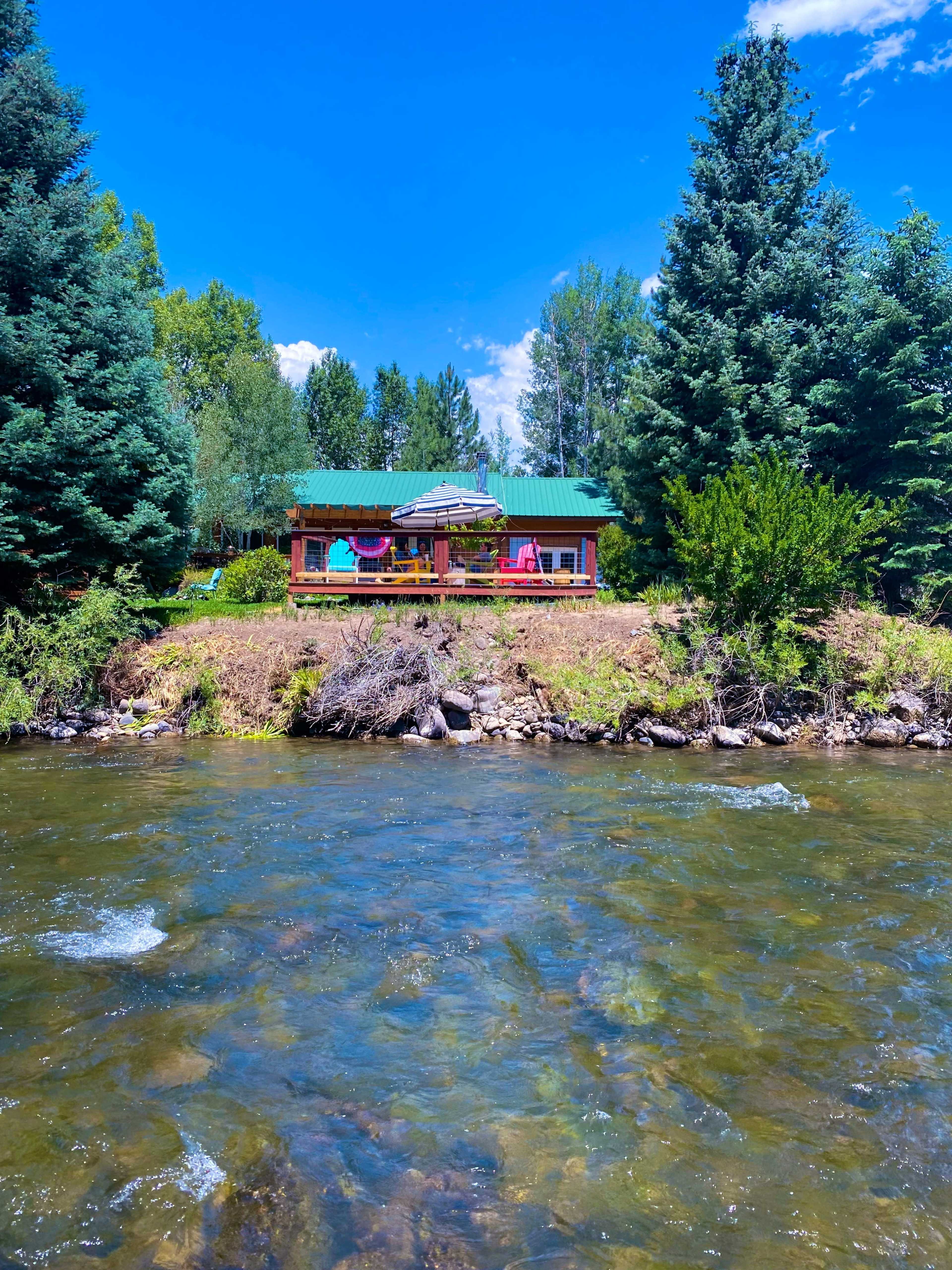 A wooden cabin with a green metal roof is situated by a river, surrounded by tall trees and colorful chairs on its deck.