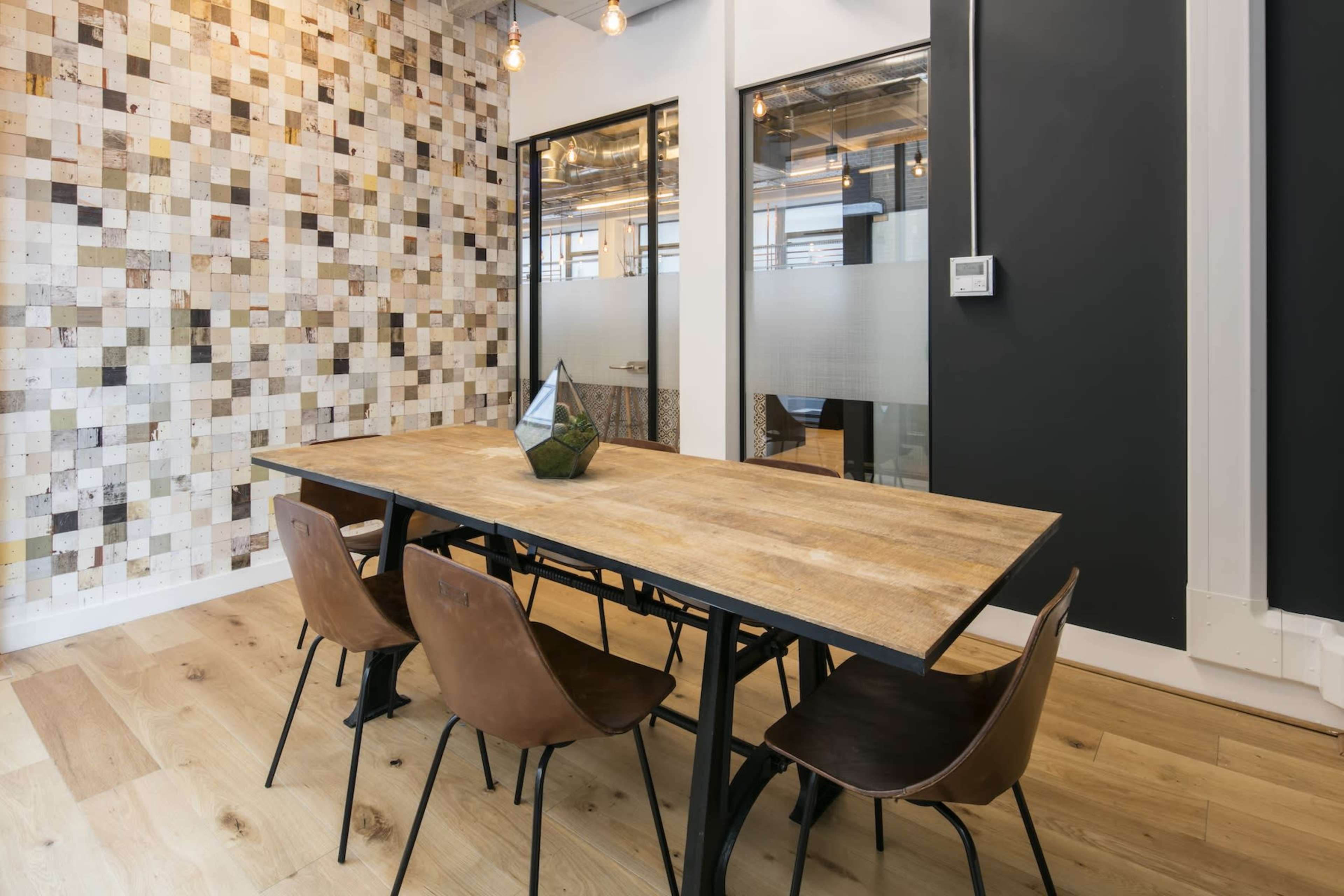 A wooden table with metal legs is set for dining in a room featuring a mosaic wall and glass-paneled office entrances.