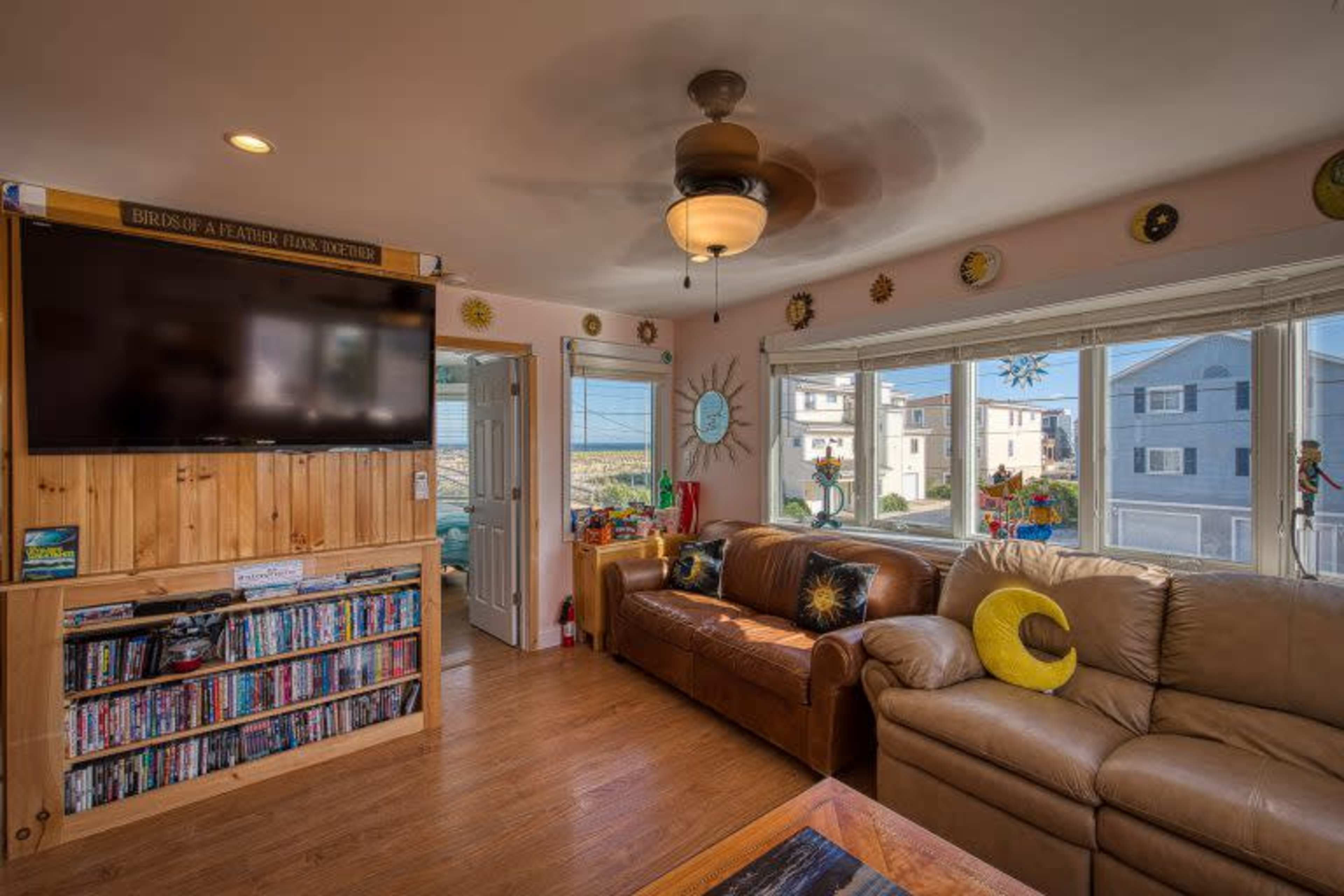 A living room with a beige couch, a large flat-screen TV mounted on a wooden wall, and windows offering a view of an outside area with other houses.