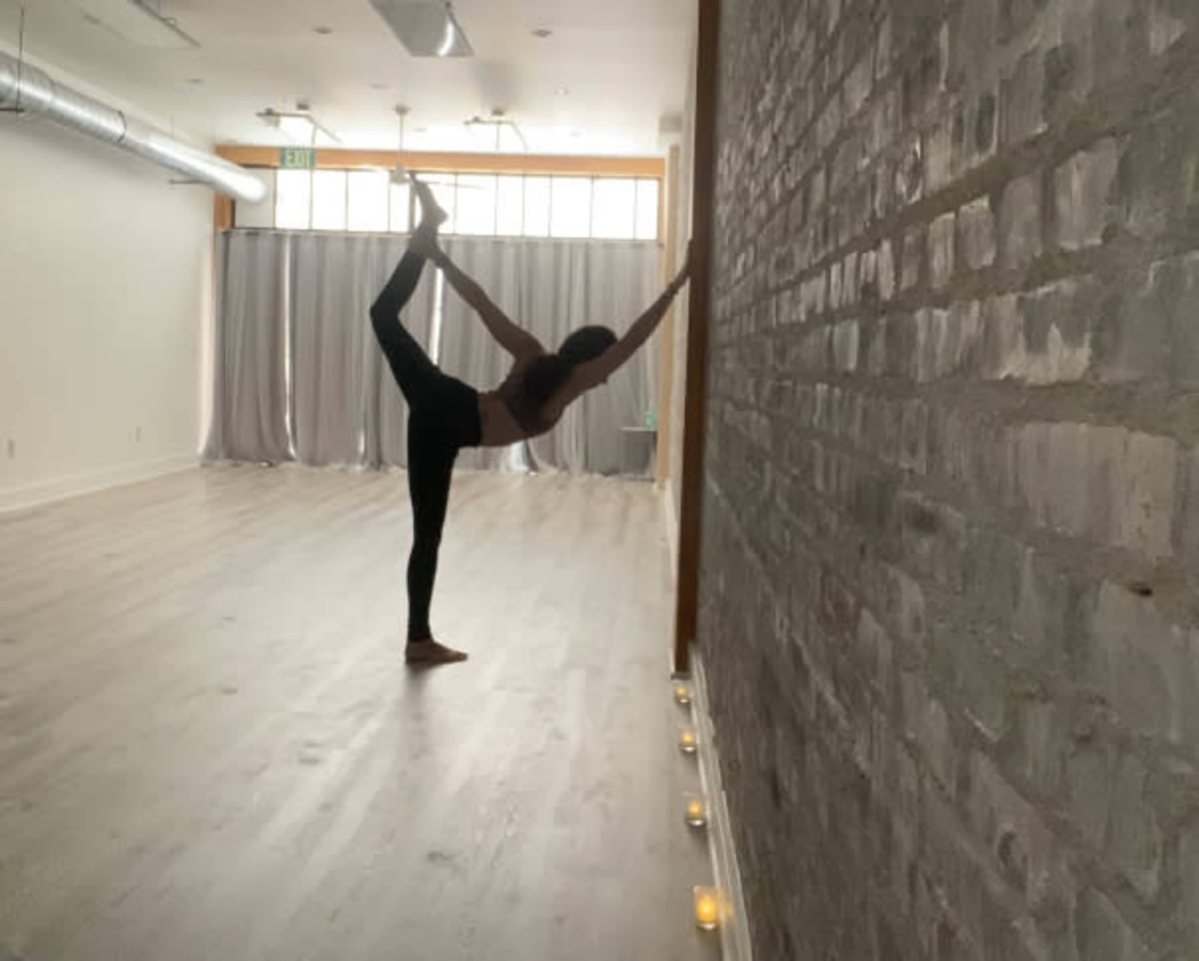 A person performs a yoga pose while balancing on one leg and reaching toward a wall in a spacious, well-lit room.