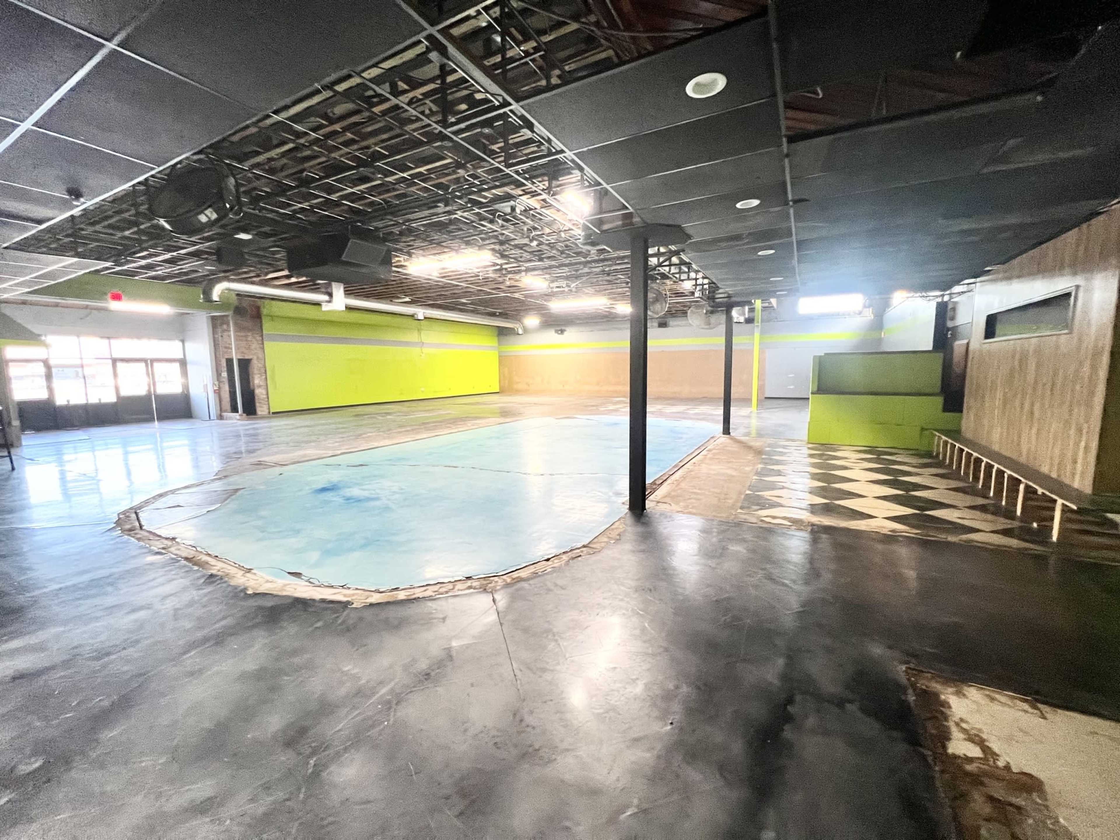 The image shows a spacious, empty room with a blue floor section and a checkered pattern in one area, surrounded by green walls and unfinished ceilings.