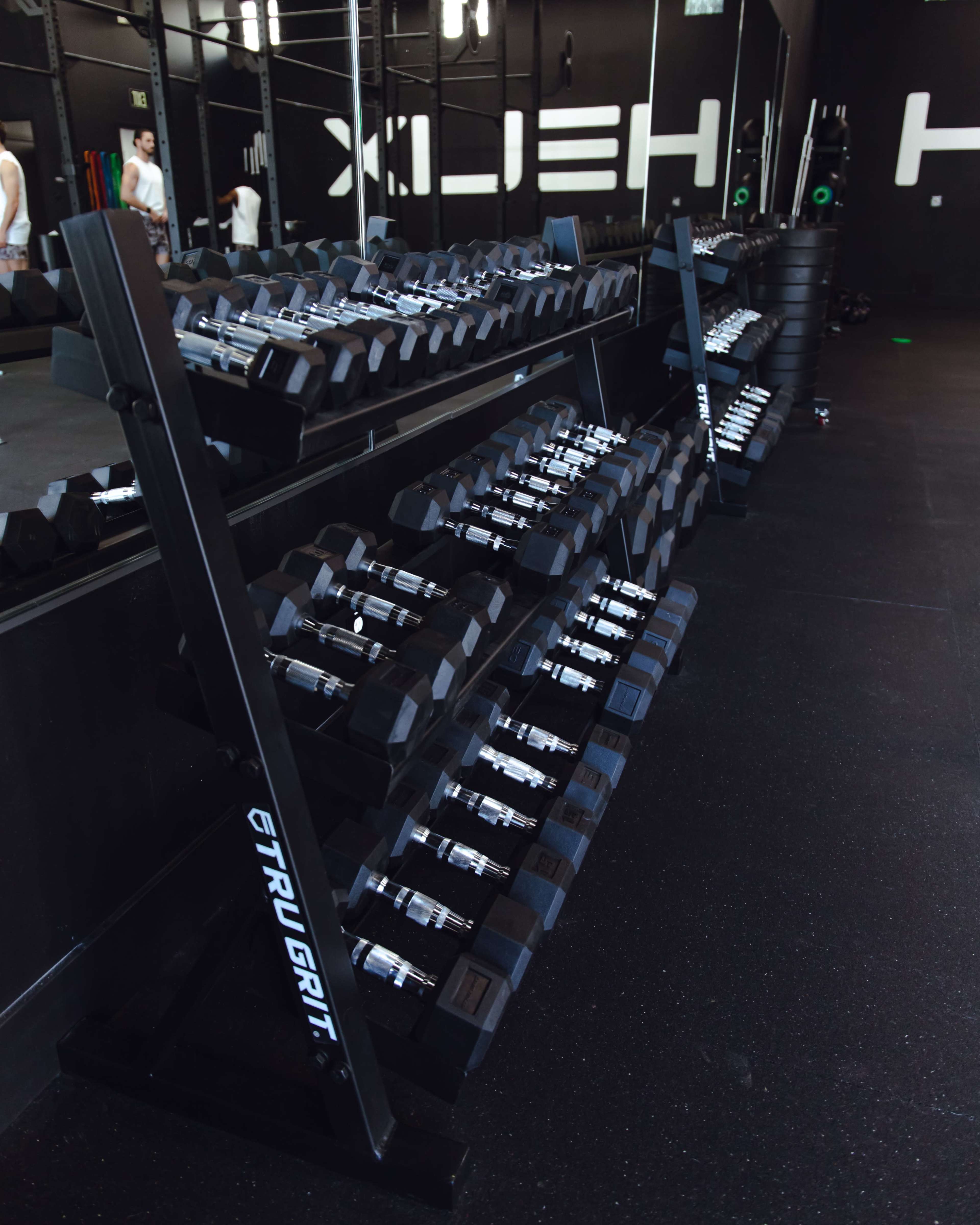 A row of dumbbells is neatly arranged on a rack in a gym with a black backdrop and fitness equipment in the background.
