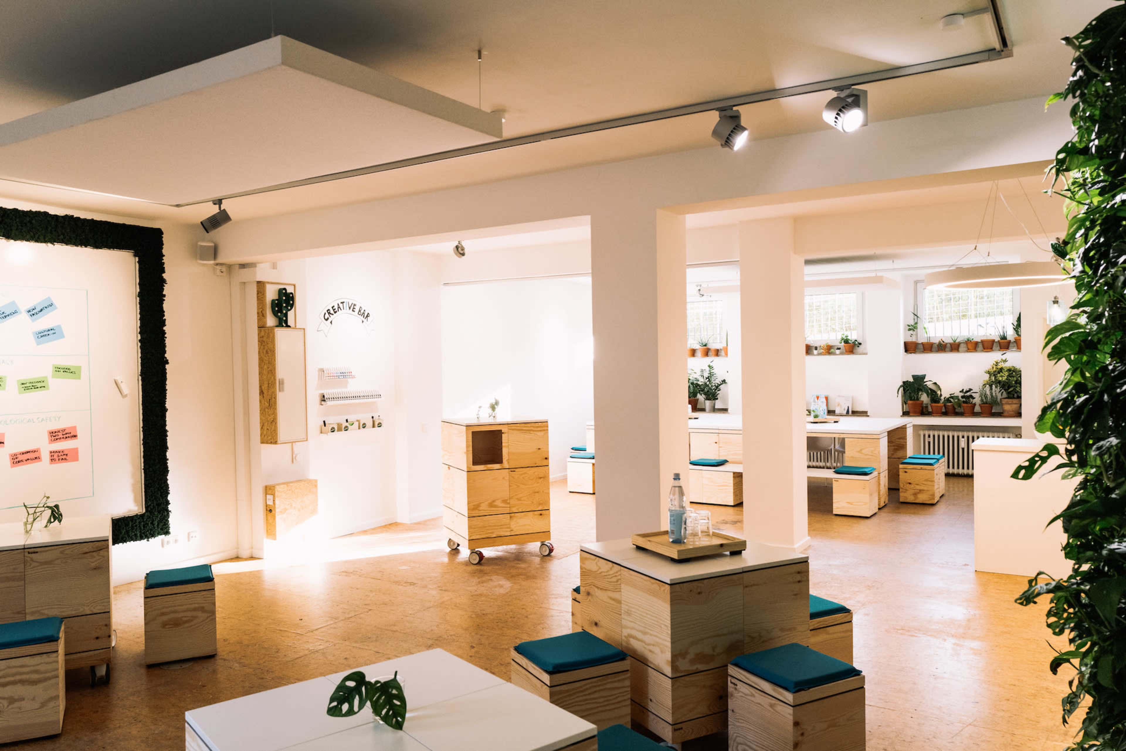 The image depicts a bright, open workspace featuring wooden boxes and tables arranged around potted plants and walls adorned with colorful notes.