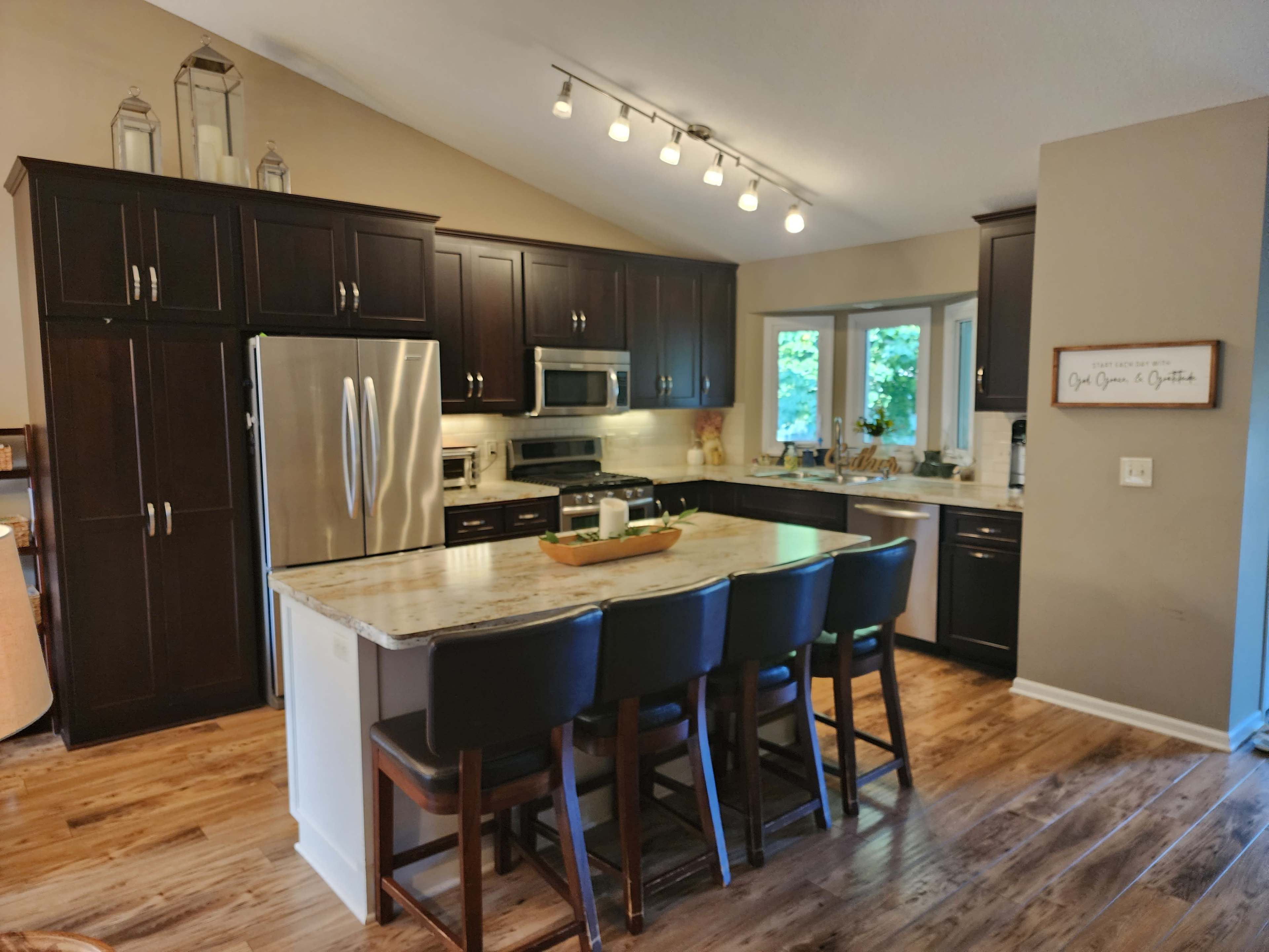 A modern kitchen features dark wood cabinetry, stainless steel appliances, a large island with seating, and natural light streaming through the windows.