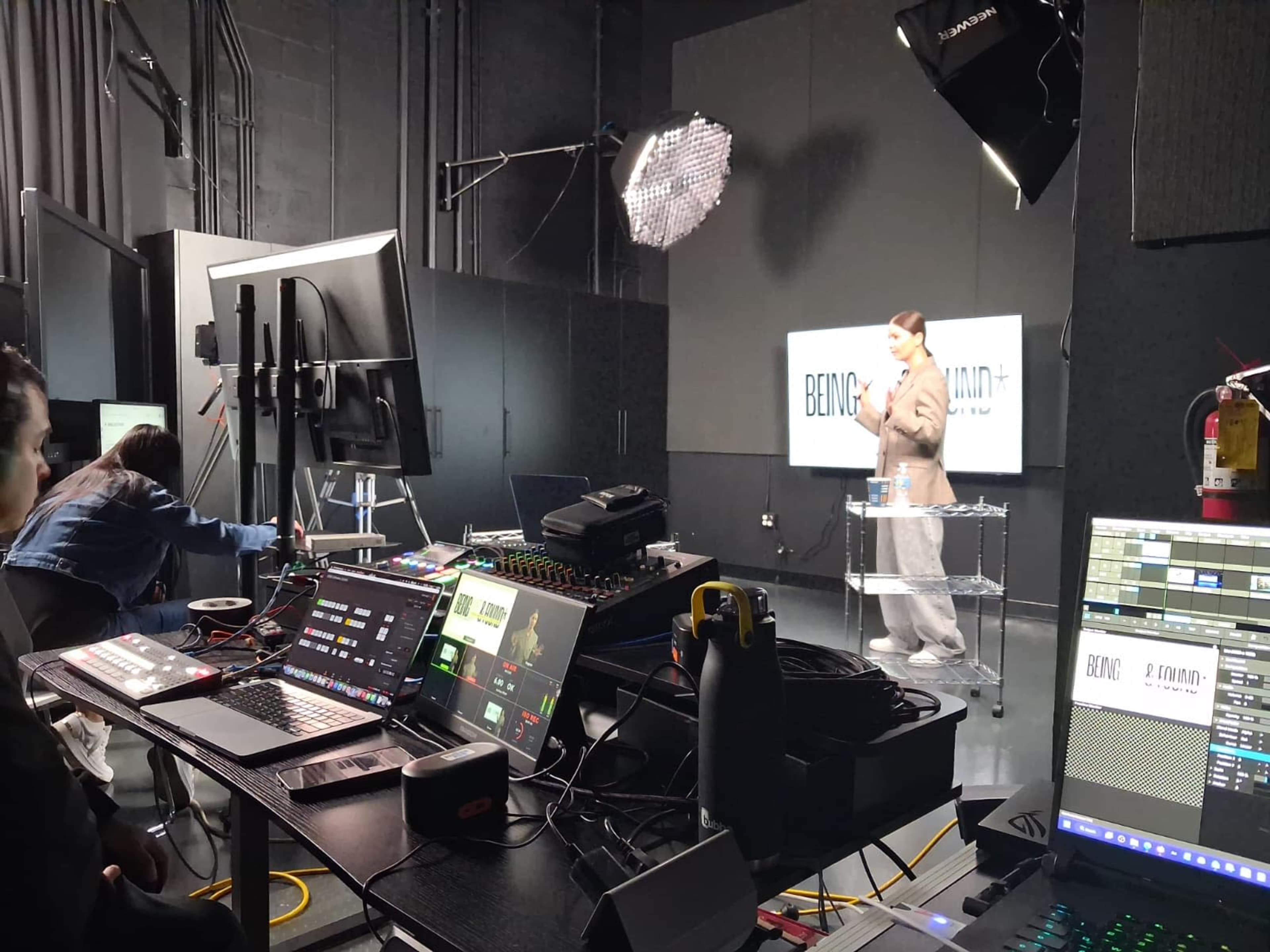 A person in formal attire is presenting in a studio setting, while a crew operates various audiovisual equipment nearby.
