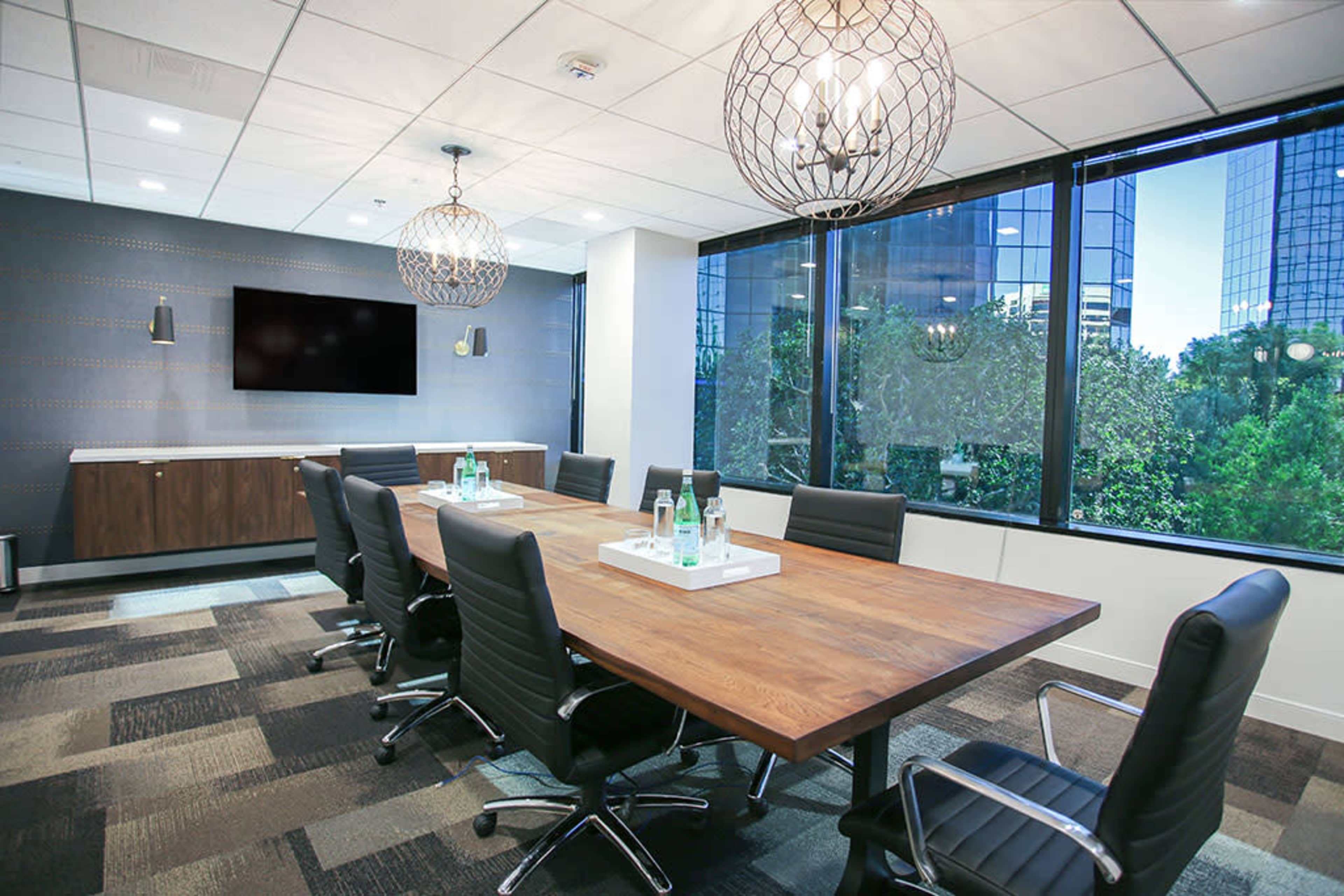 A modern conference room features a long wooden table surrounded by black chairs, with large windows providing a view of greenery outside.