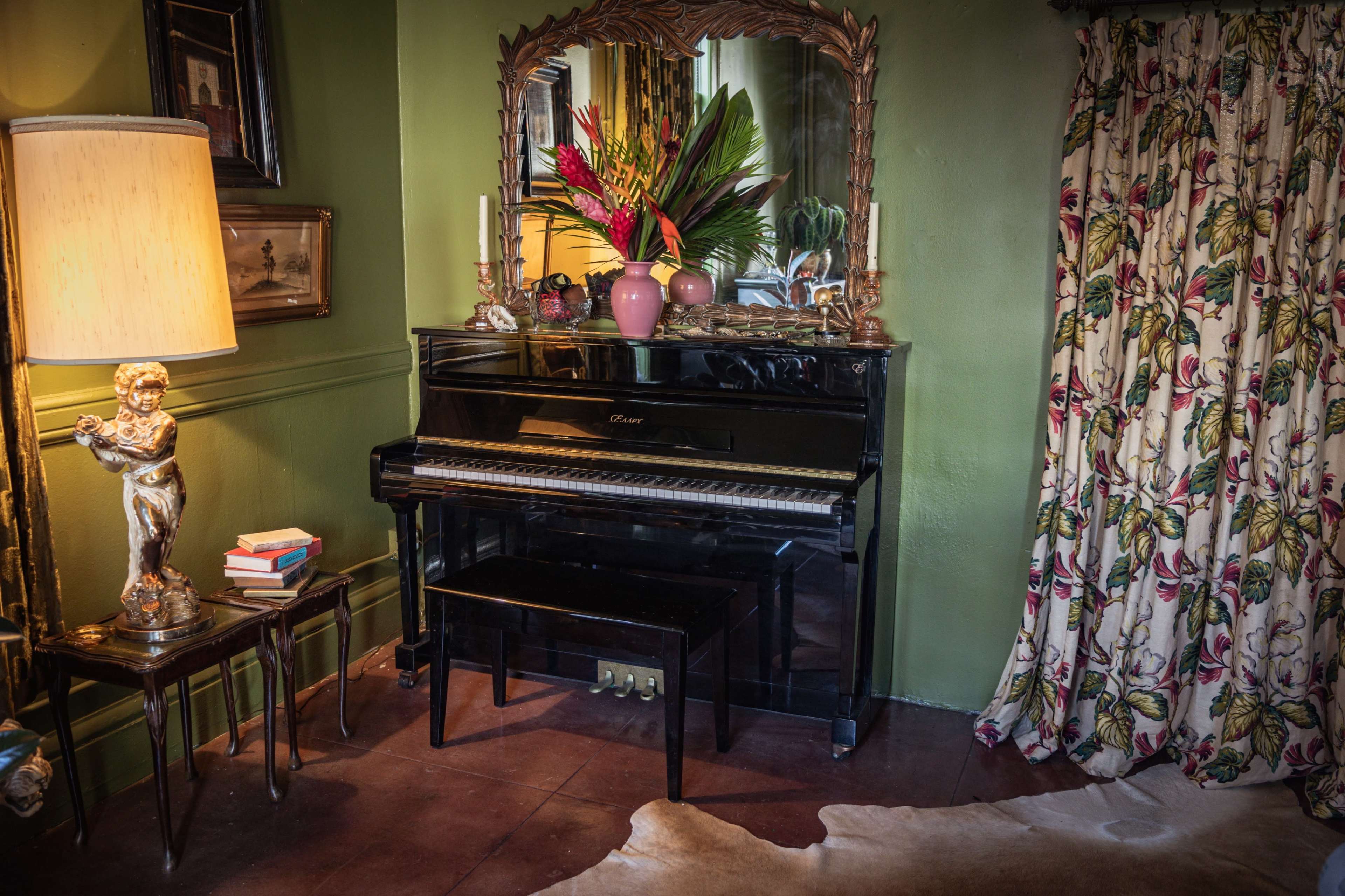 A black piano is positioned against a green wall, accompanied by a decorative mirror, a lamp, a small table with books, and a curtain featuring a floral pattern.