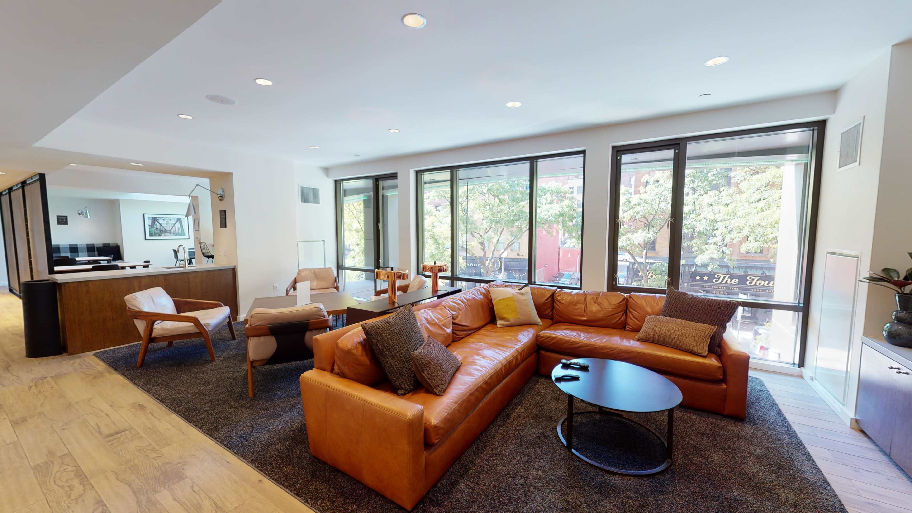 A modern living room featuring a large brown leather sectional sofa, a round black coffee table, and large windows that provide natural light and a view of outdoor greenery.