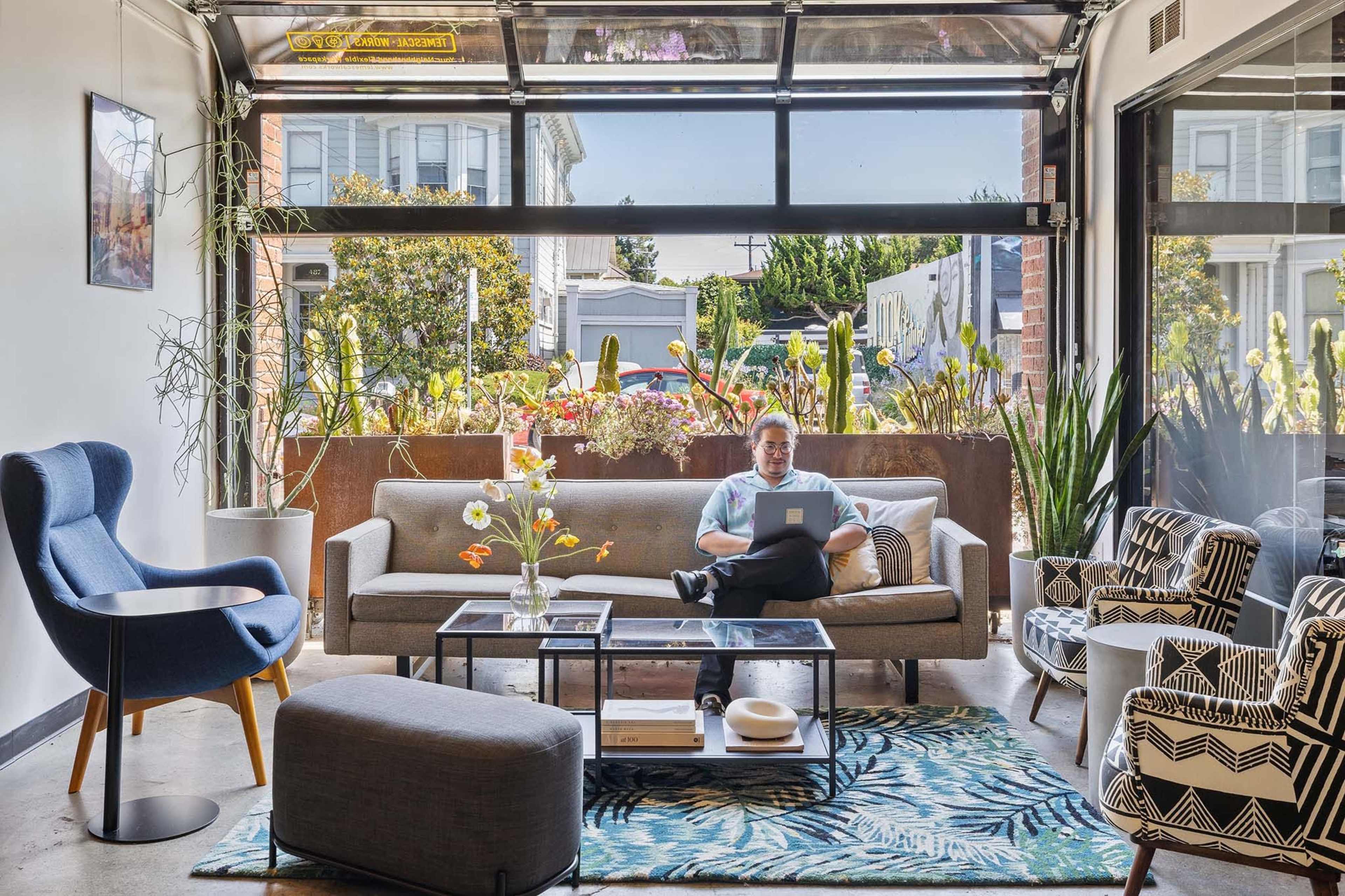 A person sits on a sofa using a laptop in a modern living room with large windows overlooking a garden.