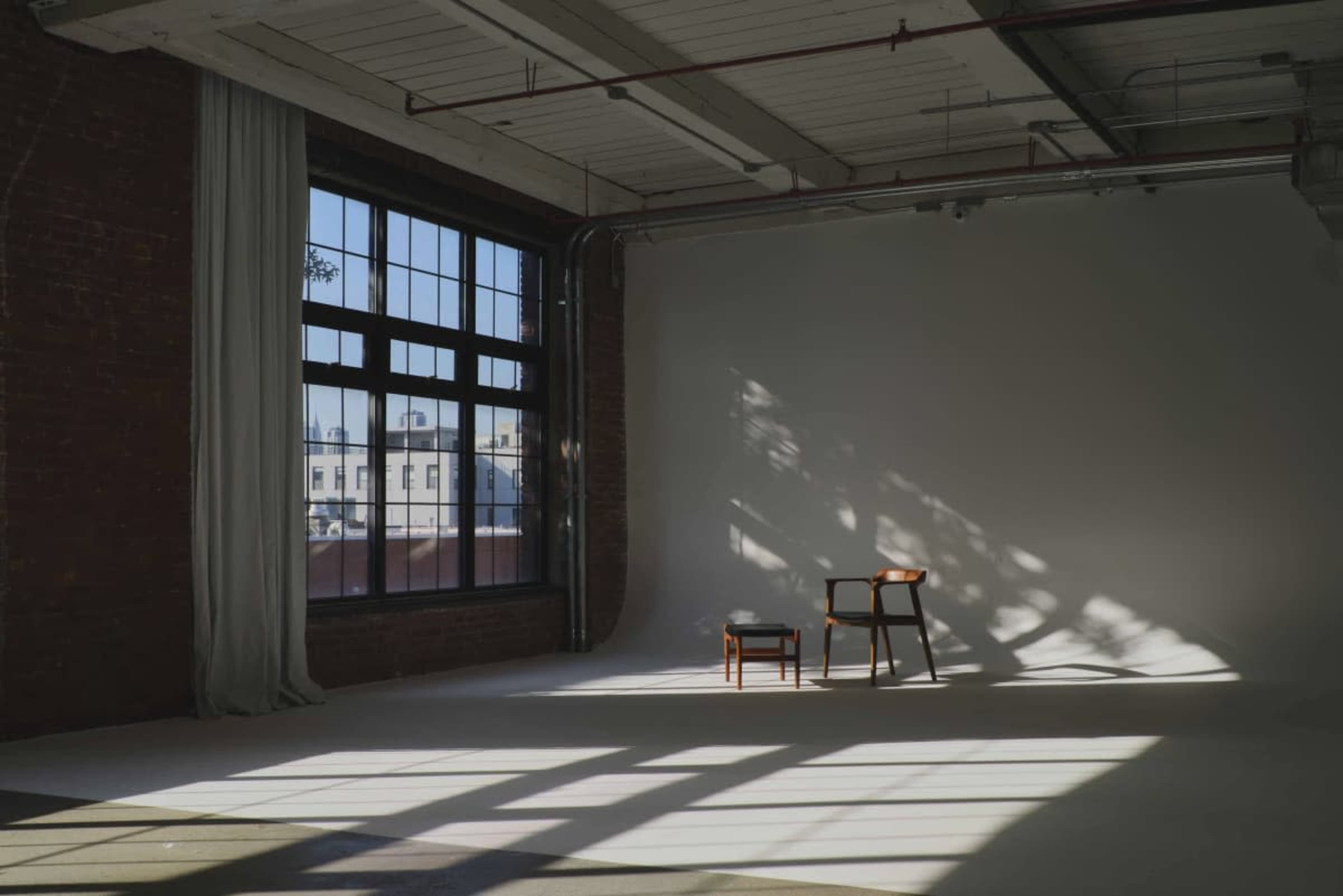A wooden chair sits alone in a bright room with large windows casting shadows on the floor.