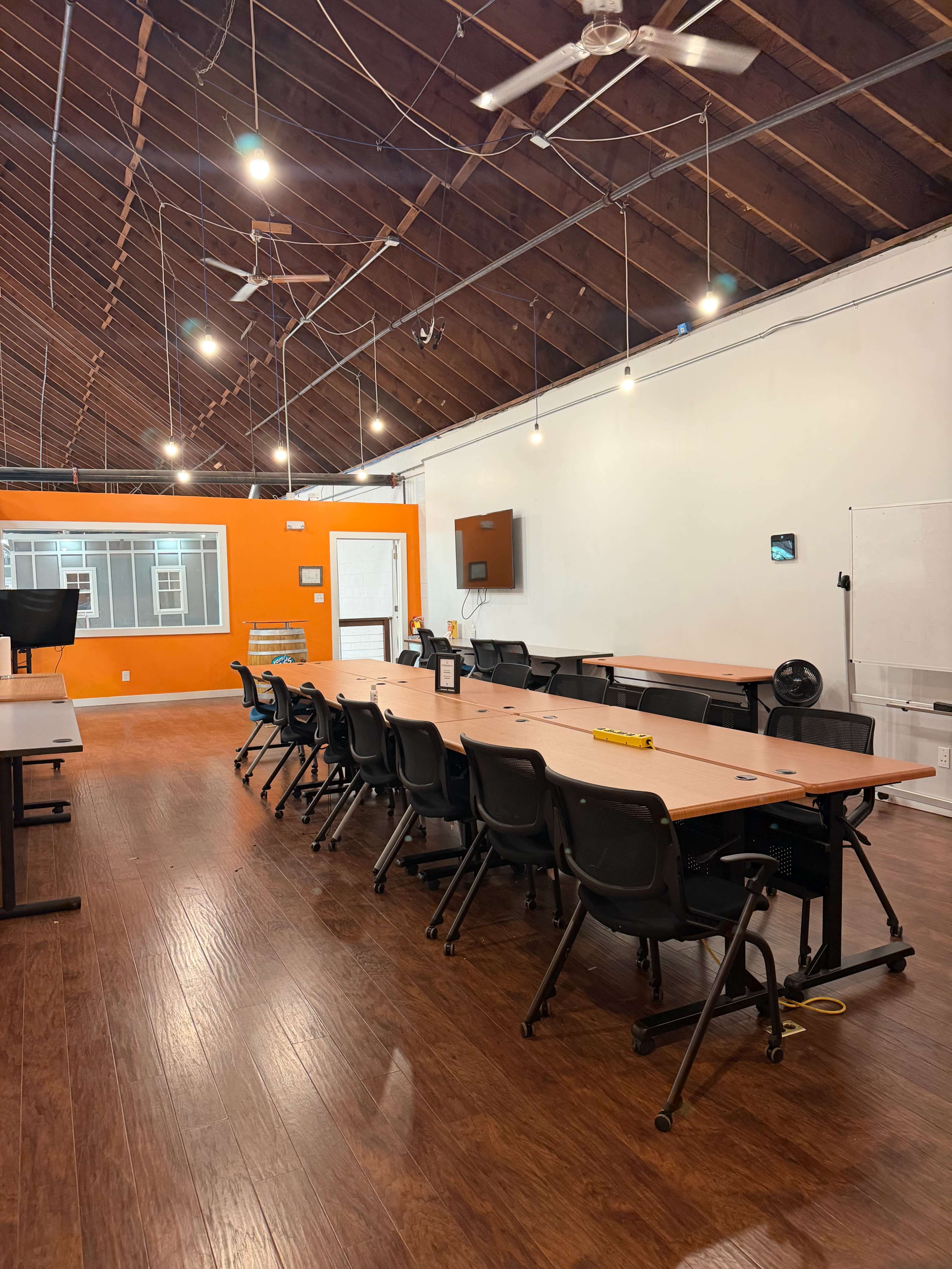 The image shows a spacious meeting room with a long table arranged with black chairs, featuring exposed wooden beams on the ceiling and an orange accent wall.