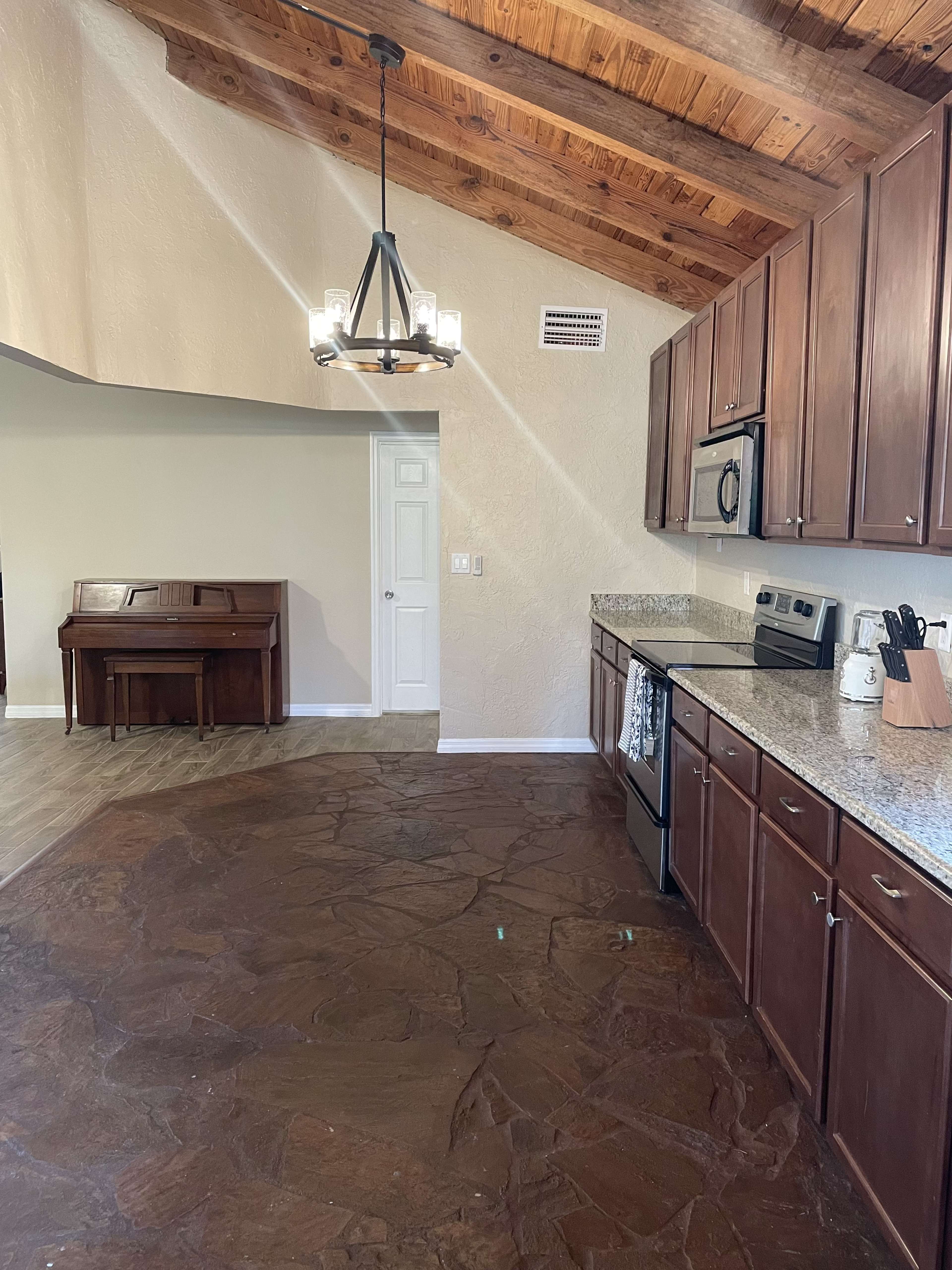 A kitchen with wooden cabinets, granite countertops, and a high ceiling with exposed beams, adjacent to a small room with a piano.