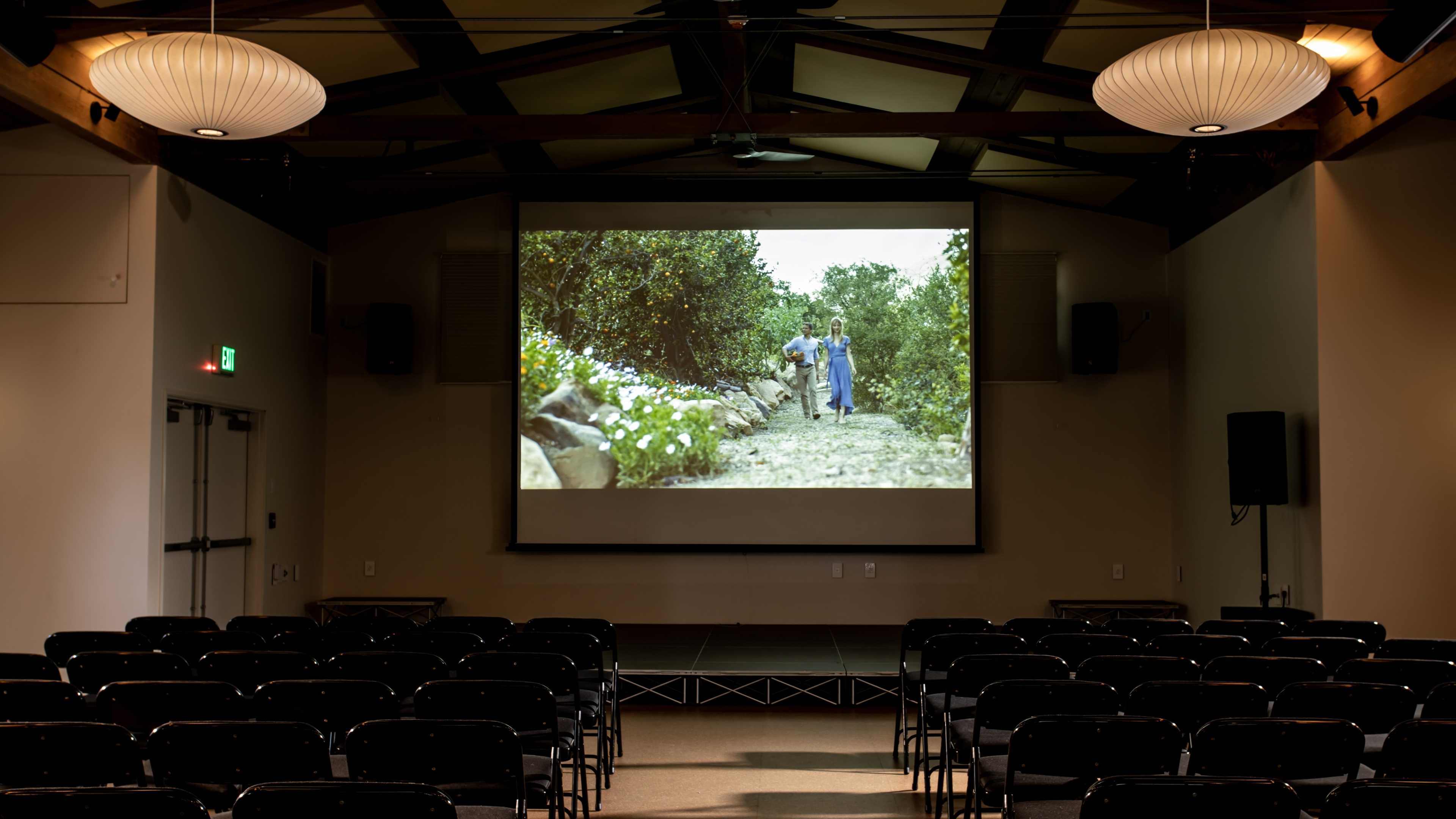 A large screen displays a film scene featuring two people walking along a path lined with greenery in an empty auditorium filled with black chairs.