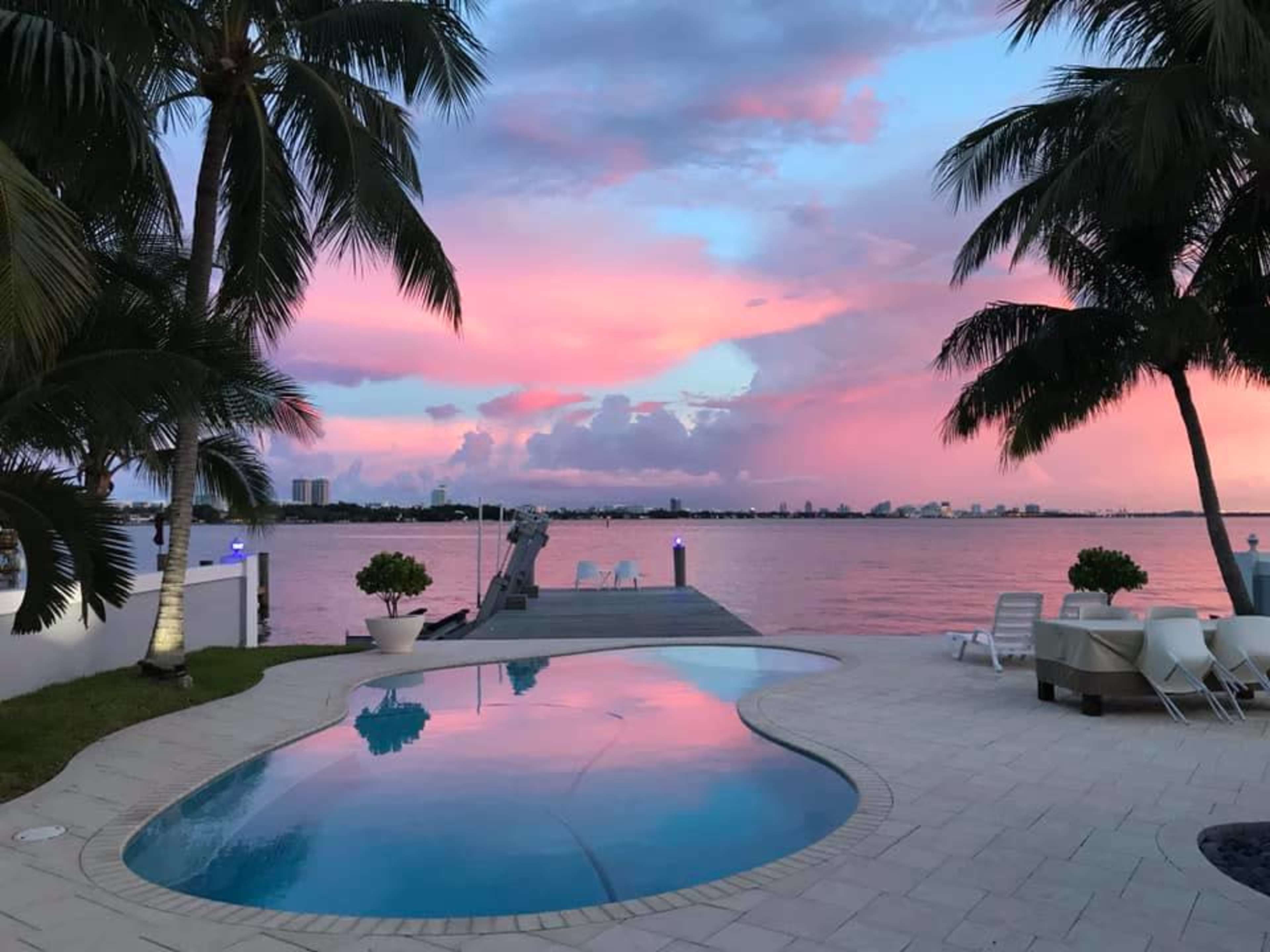 The scene shows a poolside area with palm trees and a waterfront view, featuring a vibrant pink and purple sky reflecting in the water.
