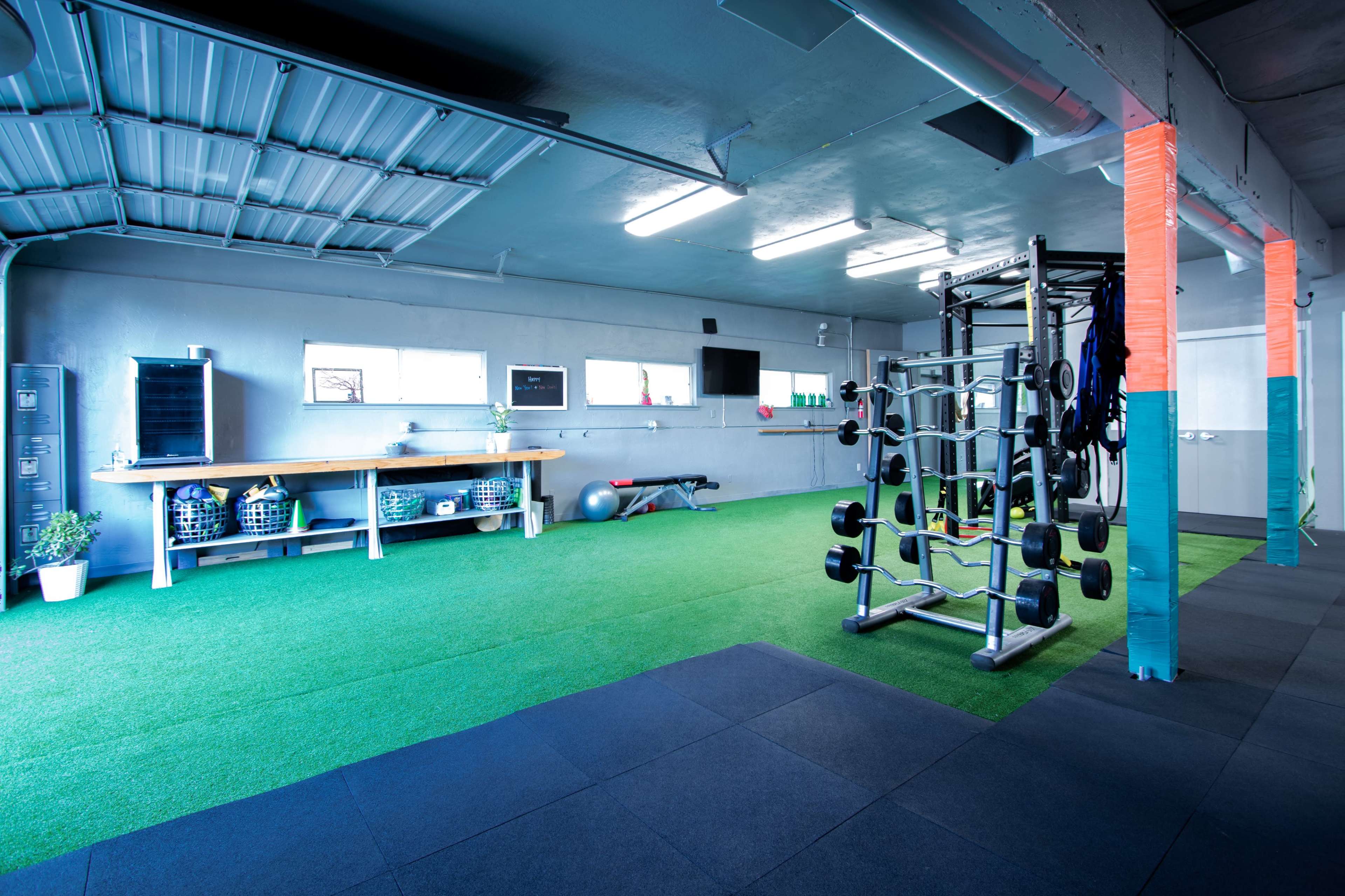 A spacious gym with green turf flooring, a weight rack, exercise equipment, and large windows providing natural light.