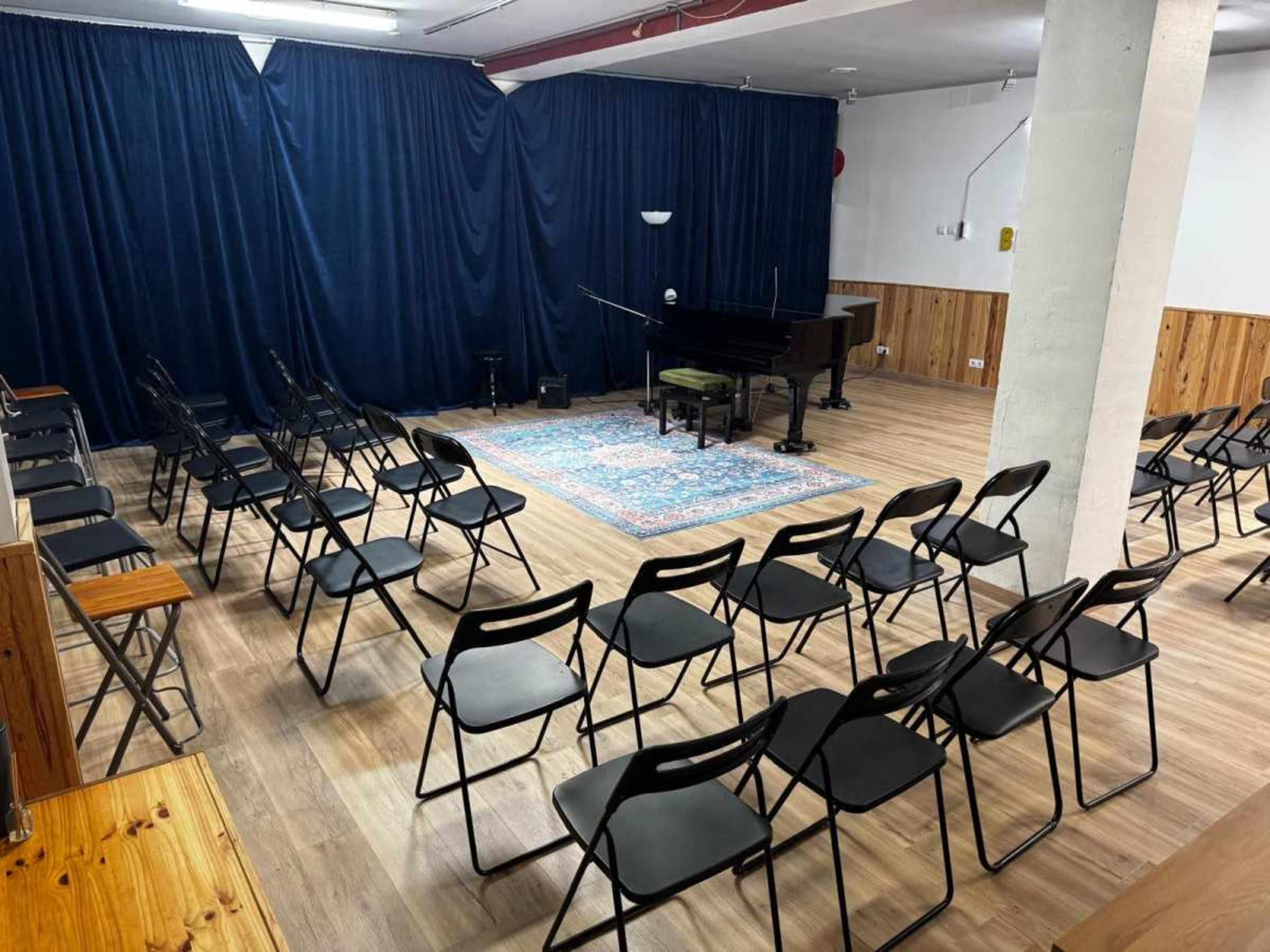 The image shows a music rehearsal space featuring a black grand piano on a rug, surrounded by rows of empty black folding chairs and blue curtains lining the walls.